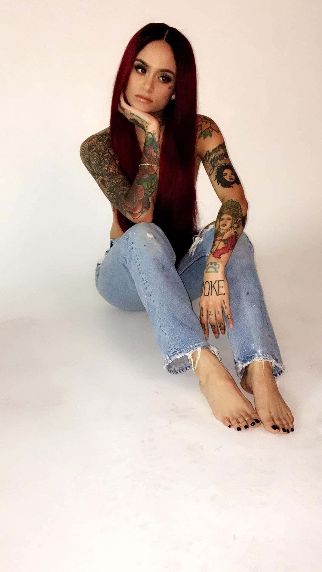 kehlani 桌布 壁紙 — 一位擁有長紅髮和全袖刺青的女性赤腳穿著破洞牛仔褲，坐在純白背景前。