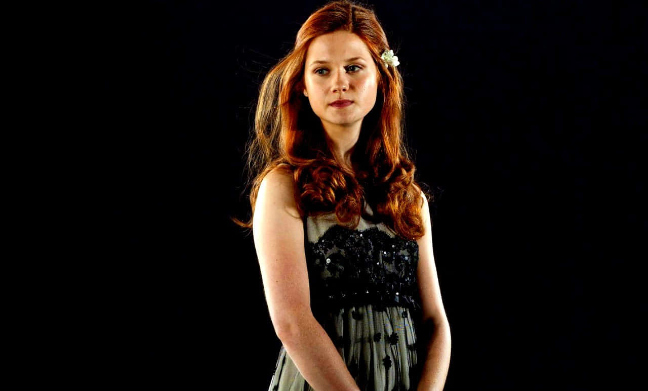Ginny Weasley 桌布 壁紙 — 一位擁有長紅髮和黑色連衣裙的女性站在深色背景前，一朵白花別在耳後。