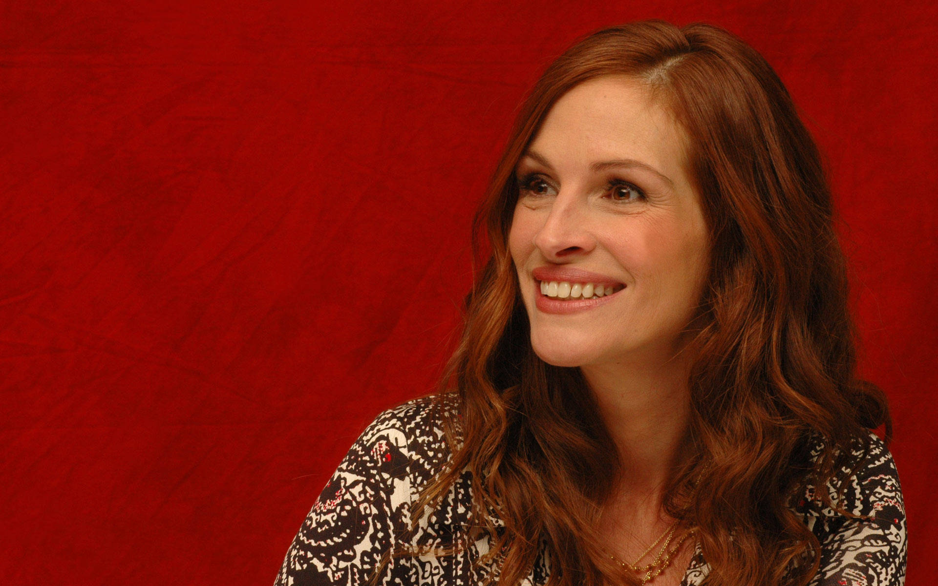 Julia Roberts 桌布 壁紙 — 一位擁有長赤褐色頭髮的女性，在帶有紋理的深紅色背景前溫暖地微笑。
