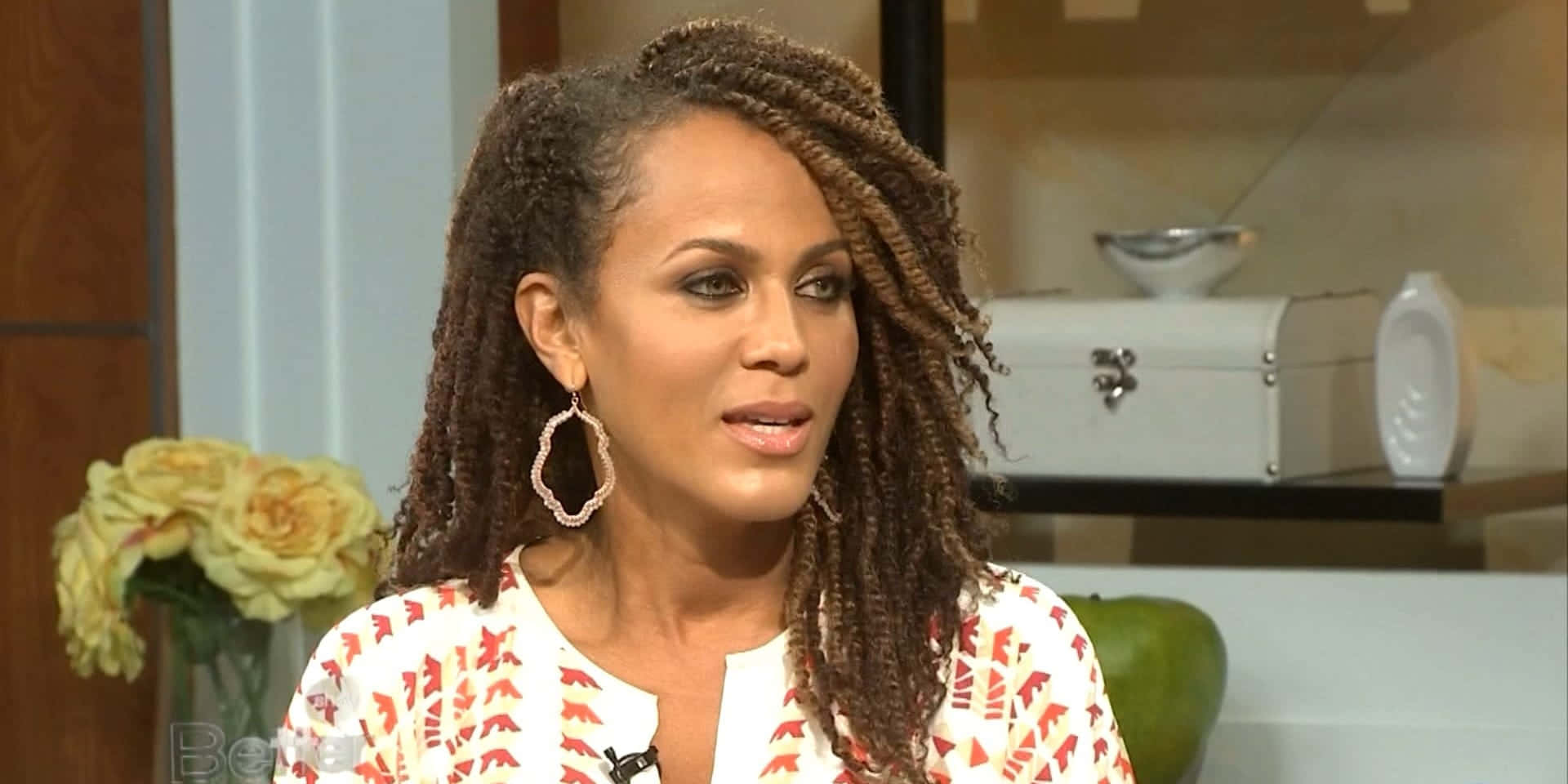 Nicole Ari Parker 桌布 壁紙 — 一位擁有長辮子和圓環耳環的女性在工作室中說話，背景有黃玫瑰和白色抽屜櫃。