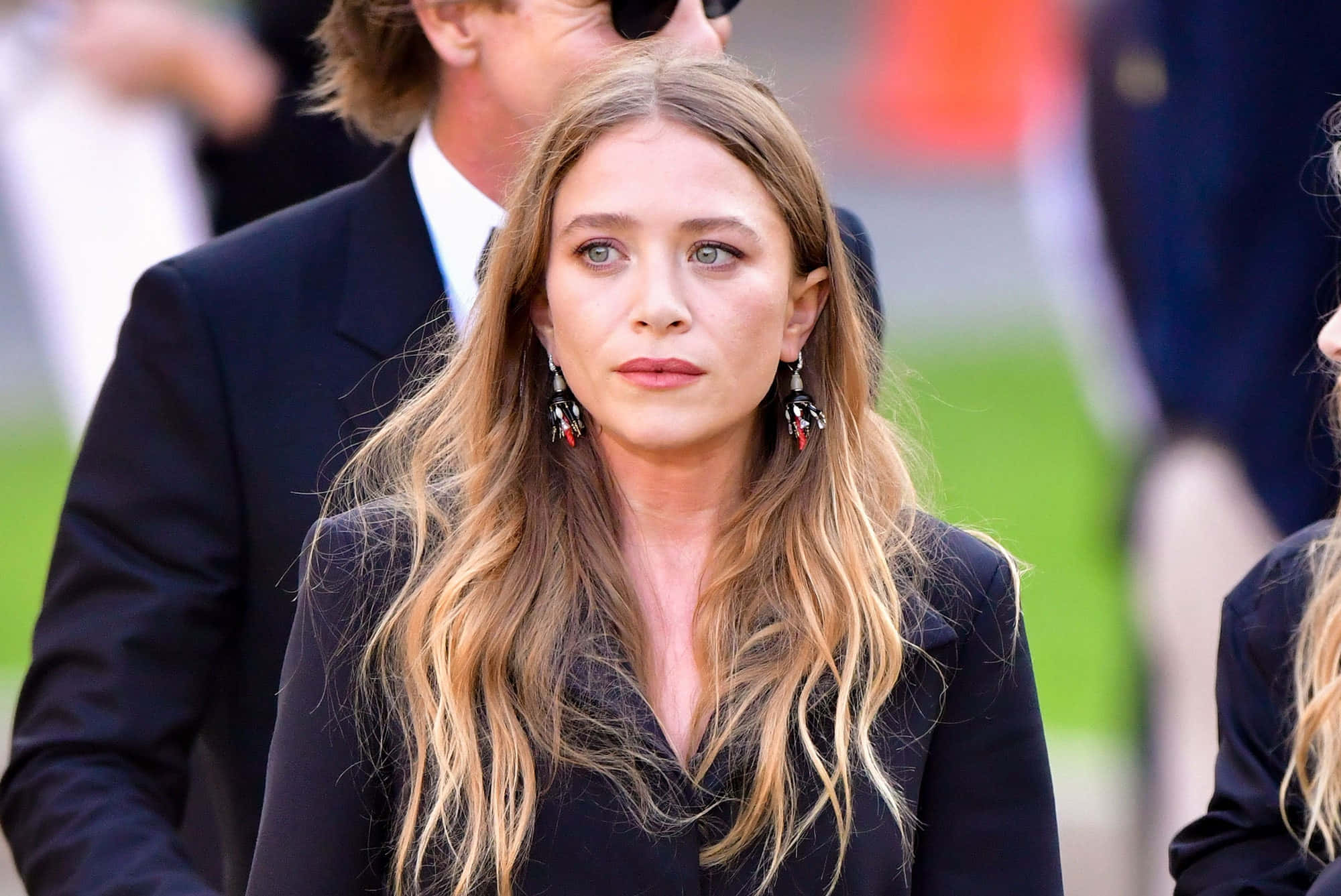 Mary Kate Olsen 桌布 壁紙 — 一位擁有長金髮和引人注目的藍眼睛的女性，穿著黑色西裝外套和垂墜耳環，站在戶外。