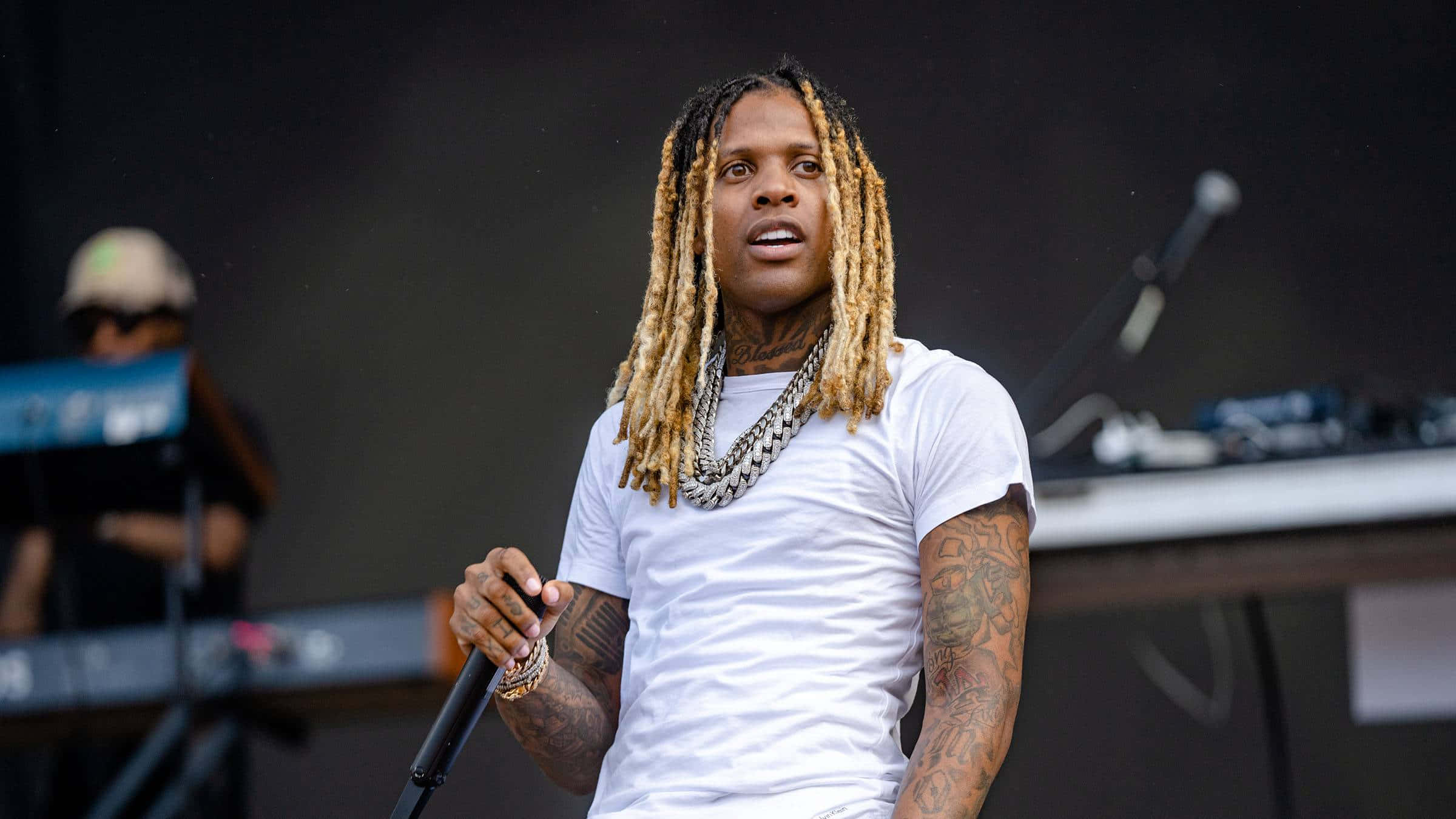 Lil Durk 桌布 壁紙 — 一位擁有長金髮辮和紋身的饒舌歌手在舞台上表演，手持麥克風，身穿白色T恤和層疊鏈條。