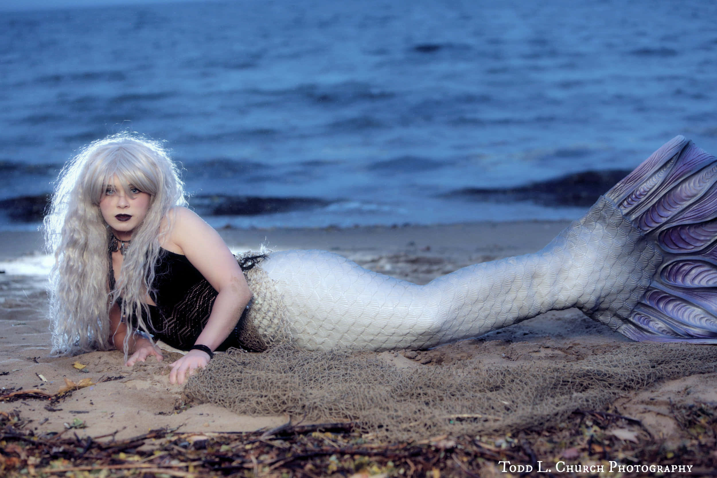 Real Mermaid 桌布 壁紙 — 一位擁有長銀色頭髮和深色妝容的人躺在沙灘上，穿著精緻的白色和紫色美人魚尾巴服裝。
