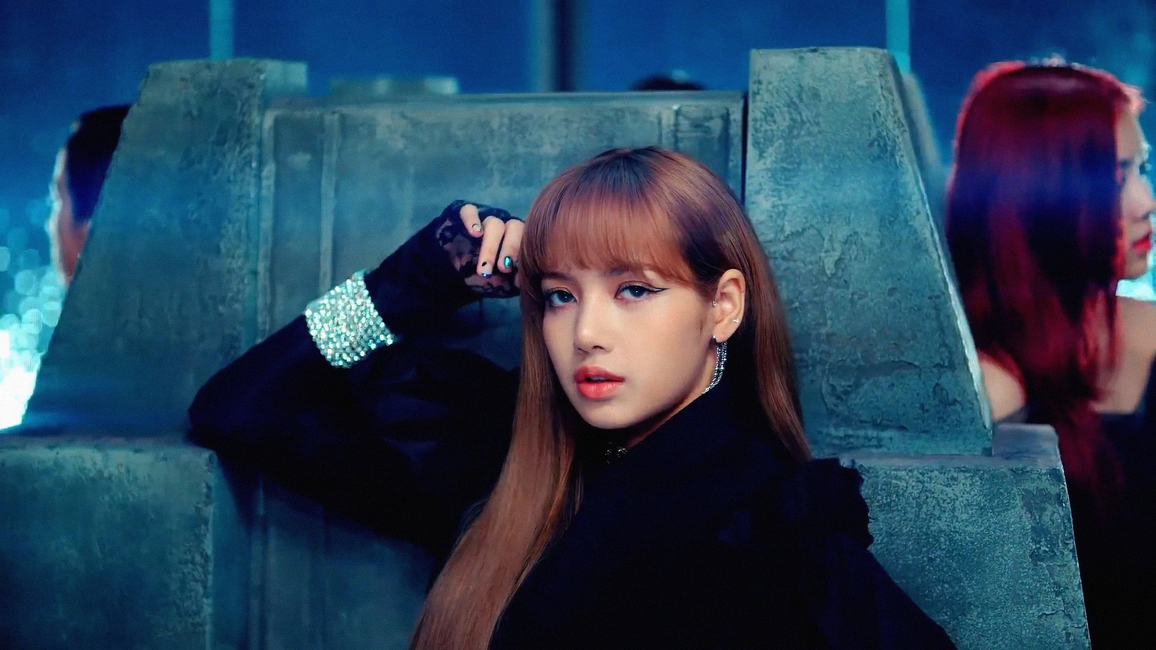 BLACKPINK 桌布 壁紙 — 一位擁有長長棕色頭髮和濃妝的女性在黑暗的未來主義場景中自信地擺姿勢，場景帶有藍色燈光和石頭狀結構。