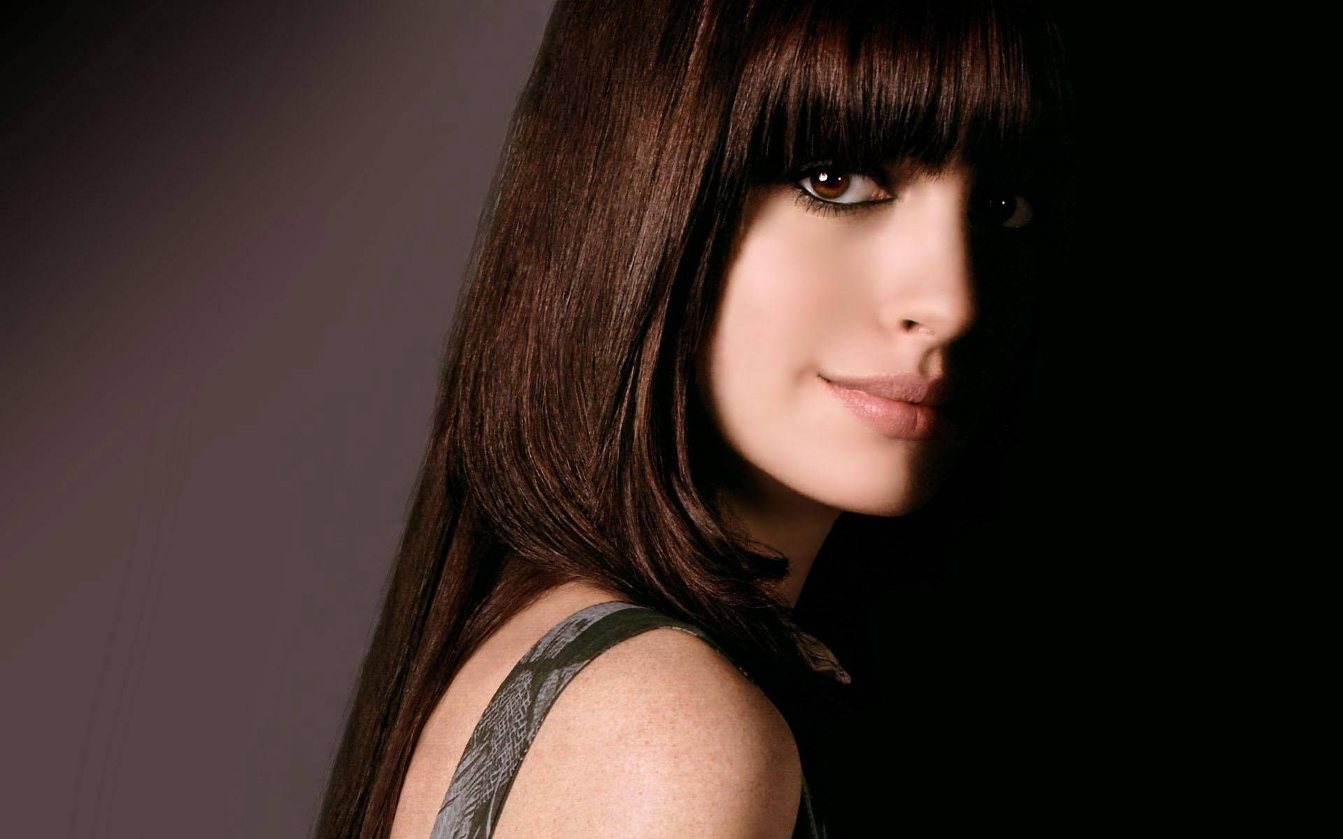 Anne Hathaway 桌布 壁紙 — 一位擁有長長棕色頭髮和瀏海的女性，在昏暗的攝影棚中，在戲劇性的燈光下溫柔地凝視。