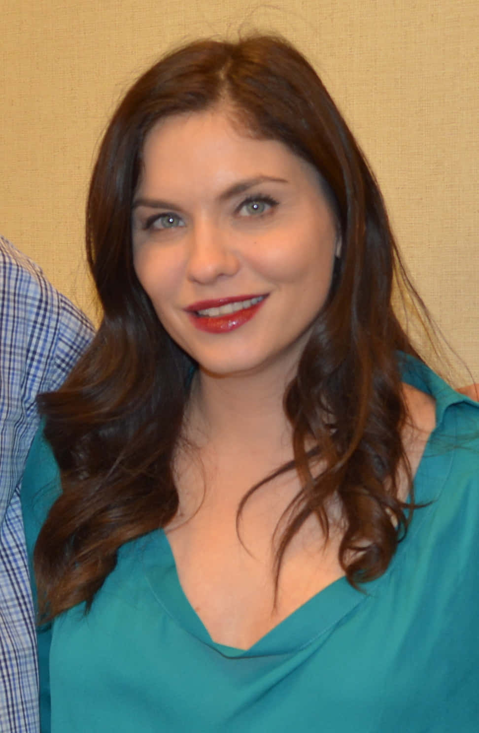 jodi lyn o'keefe 桌布 壁紙 — 一位擁有長長棕色頭髮和紅色唇膏的女性，穿著藍綠色上衣，在米色紋理牆壁前溫暖地微笑。