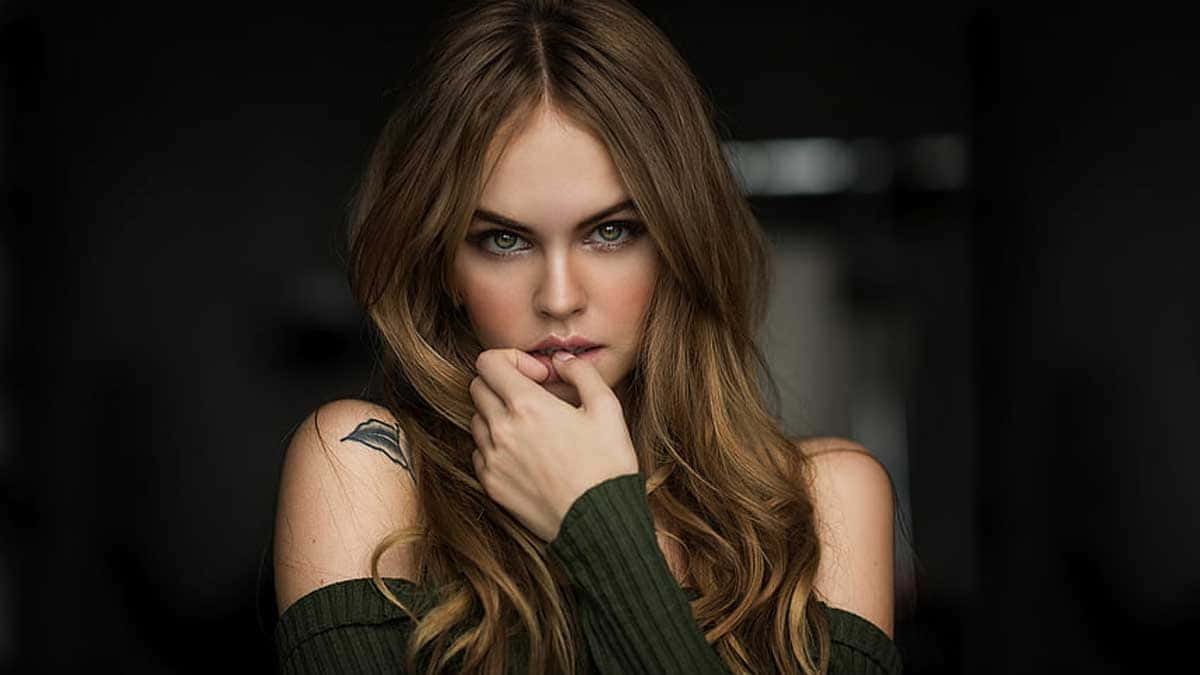 Celebrity 桌布 壁紙 — 一位擁有長長棕色頭髮和紋身的女性，在深色背景下觸摸嘴唇，凝視著強烈的目光。
