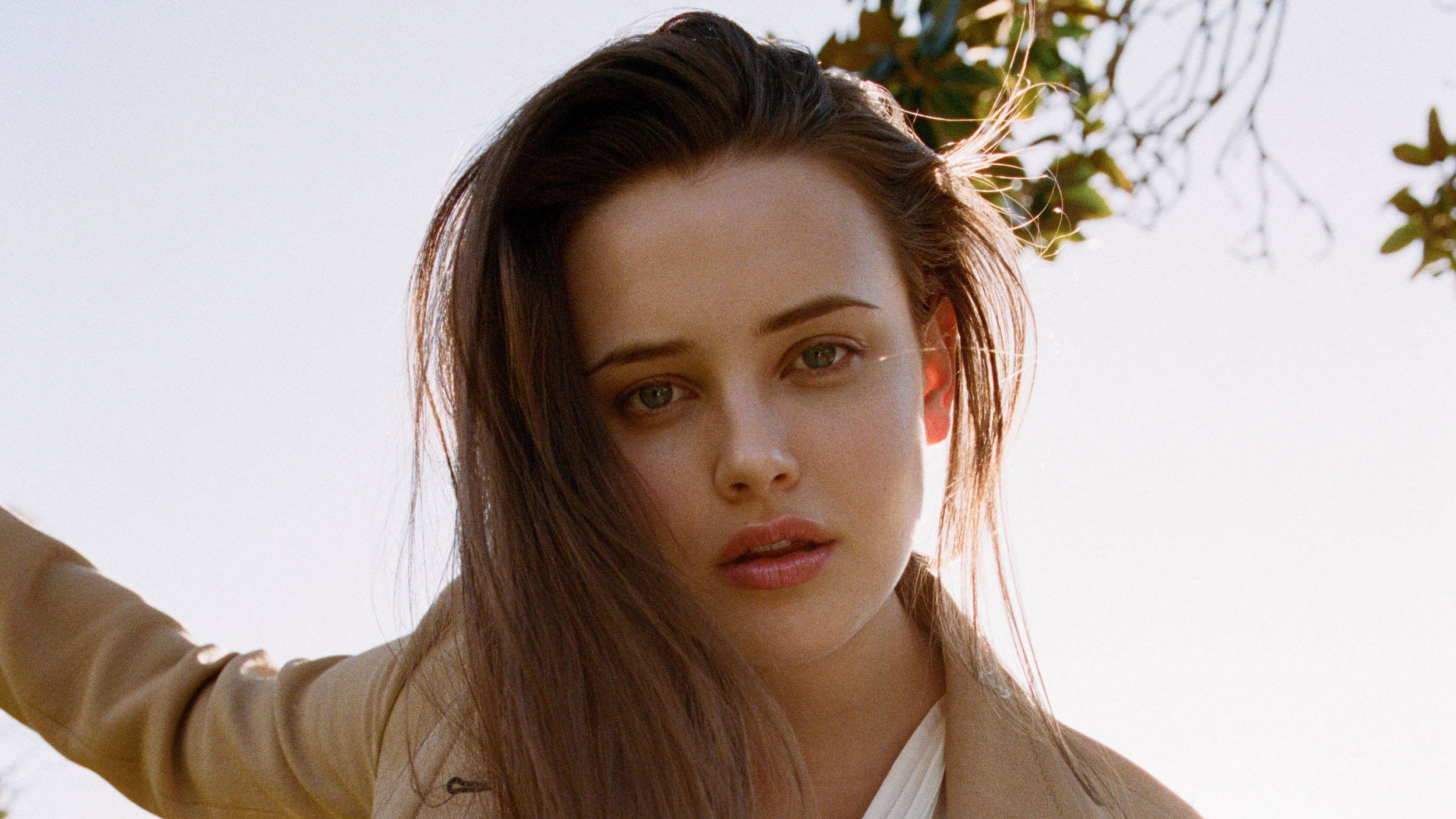 Katherine Langford 桌布 壁紙 — 一位擁有長長棕色頭髮和綠色眼睛的女性在明亮的陽光下，以樹枝為背景，直視鏡頭。