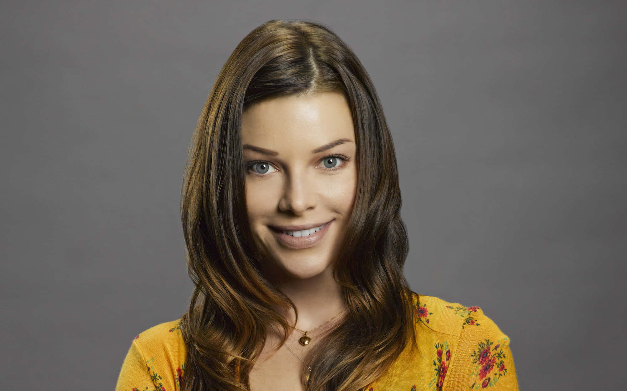 Lauren German 桌布 壁紙 — 一位擁有長長棕色頭髮和藍色眼睛的女性，在灰色背景下穿著黃色花卉上衣，溫暖地微笑。