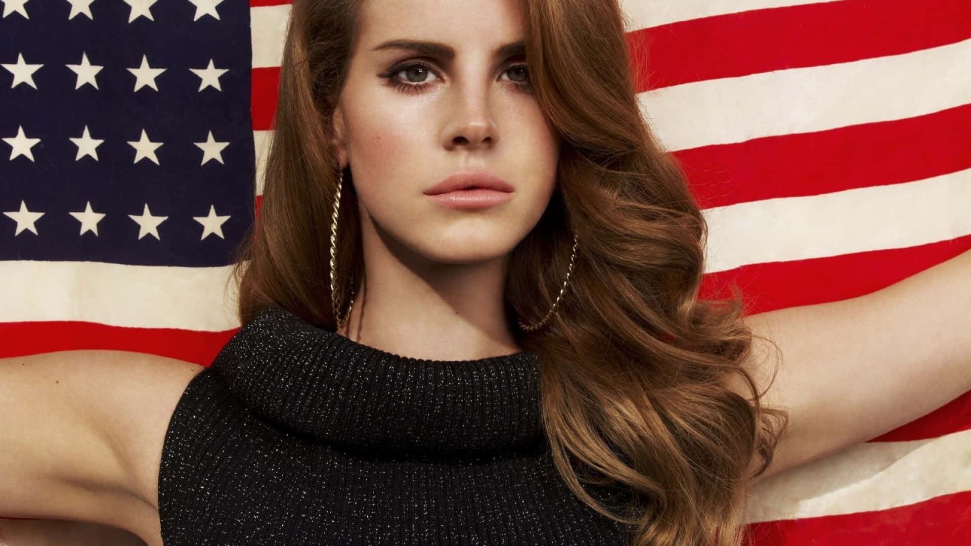 lana del rey 桌布 壁紙 — 一位擁有長長棕色頭髮和金色圓環耳環的女性，穿著黑色針織上衣，在大型美國國旗背景前自信地擺姿勢。