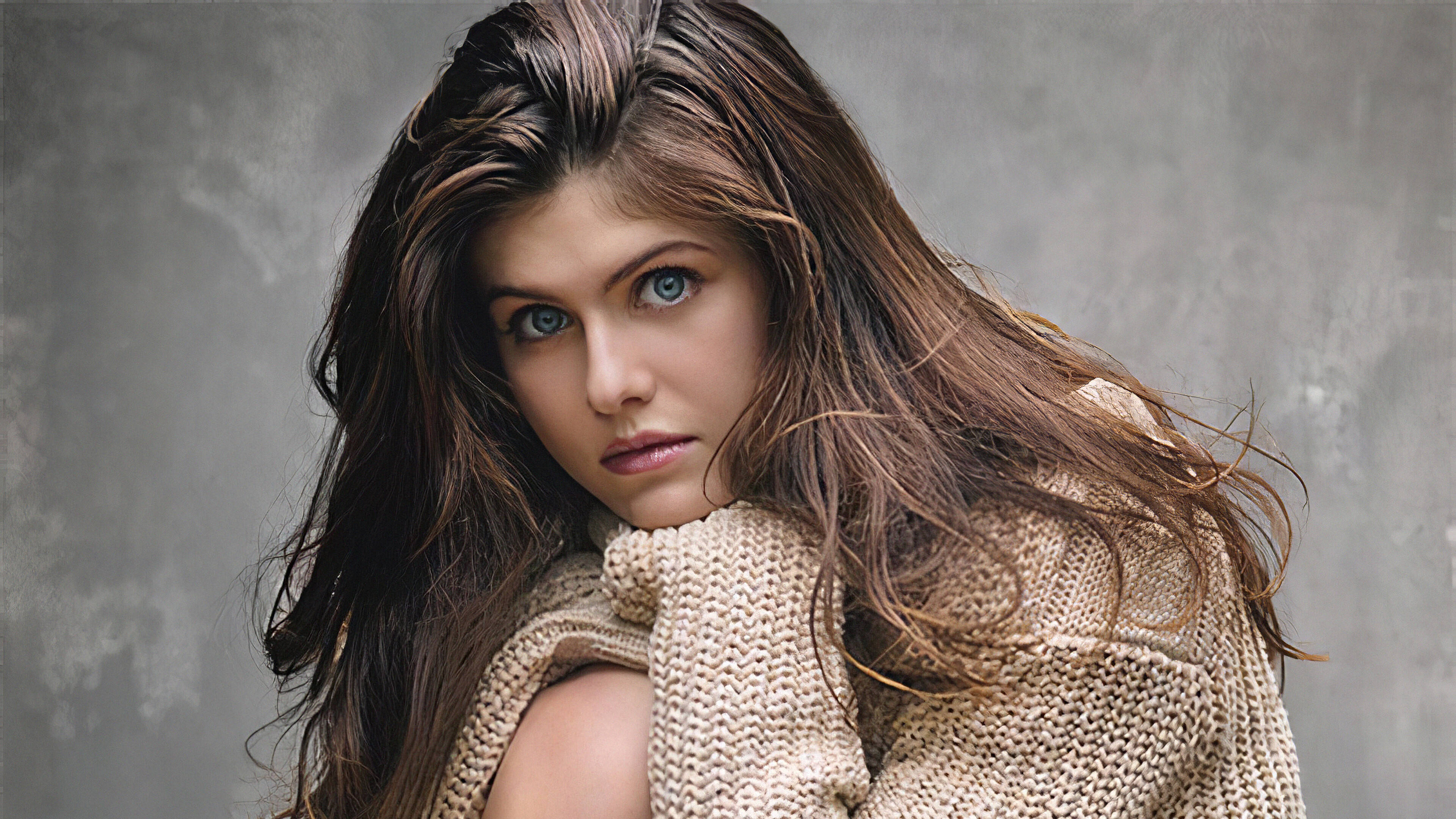Alexandra Daddario 4K 桌布 壁紙 — 一位擁有長長棕色頭髮和銳利藍色眼睛的女性，穿著米色針織毛衣，在紋理灰色牆壁前凝視著。