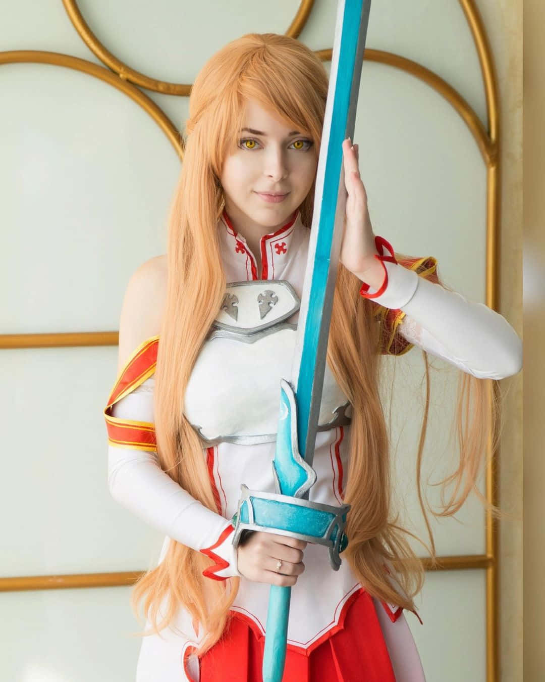 Sword Art Online Cosplay 桌布 壁紙 — 一位擁有長長橙色頭髮的角色扮演者，身穿白色、紅色和金色的服裝，在金色拱形背景前手持一把發光的藍色劍。