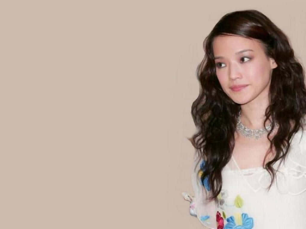 Shu Qi 桌布 壁紙 — 一位擁有長長波浪深色頭髮、身穿花卉上衣的女性，在純米色背景前若有所思地凝視著。