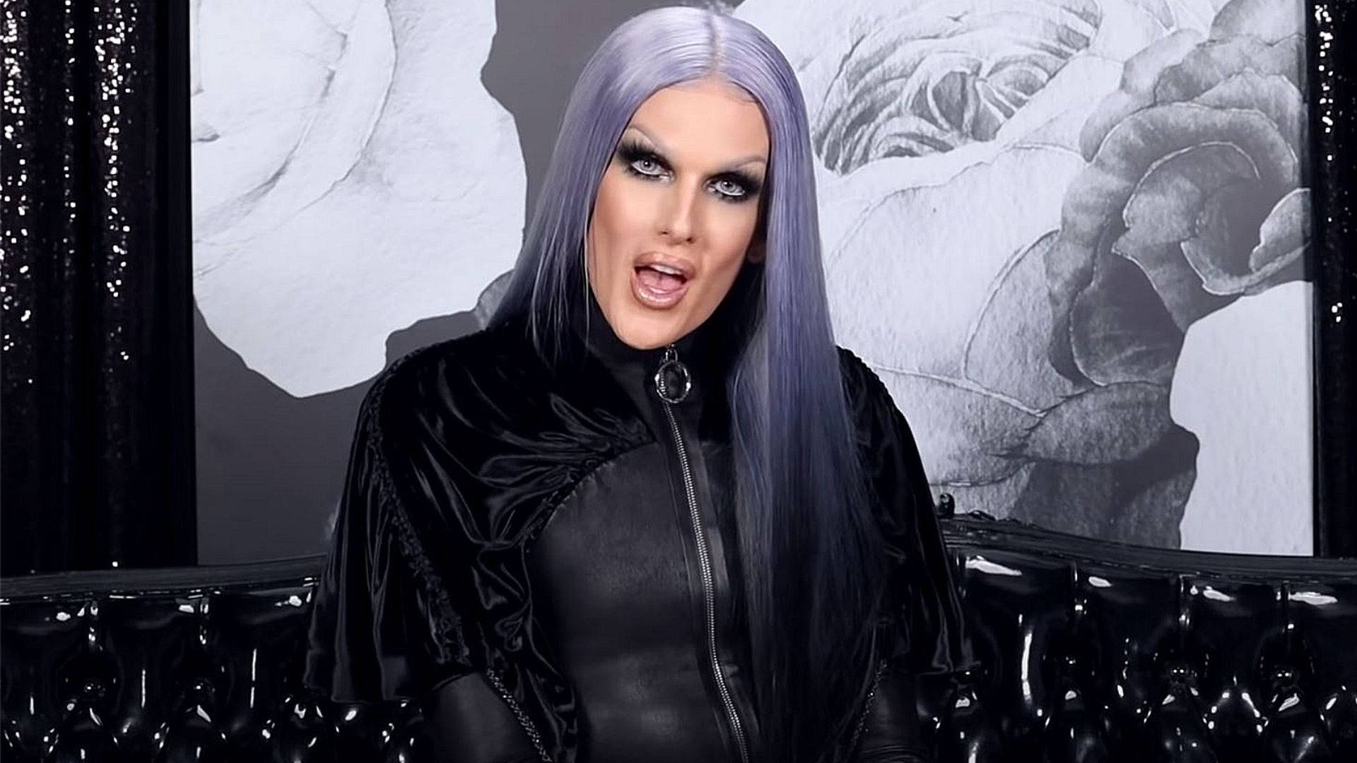 Jeffree Star 桌布 壁紙 — 一位擁有長長薰衣草色頭髮和戲劇化妝容的人，穿著黑色天鵝絨服裝，背景是單色花卉圖案。