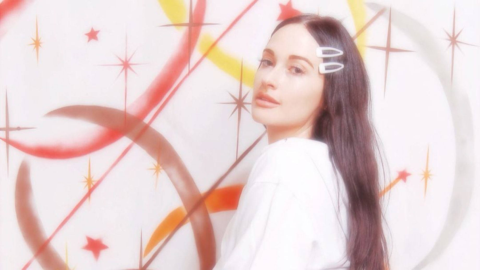 Kacey Musgraves 桌布 壁紙 — 一位擁有長長黑髮和白色夾子的女性，在充滿活力的紅、橙、黃色星星和漩渦的抽象背景前溫柔地凝視。