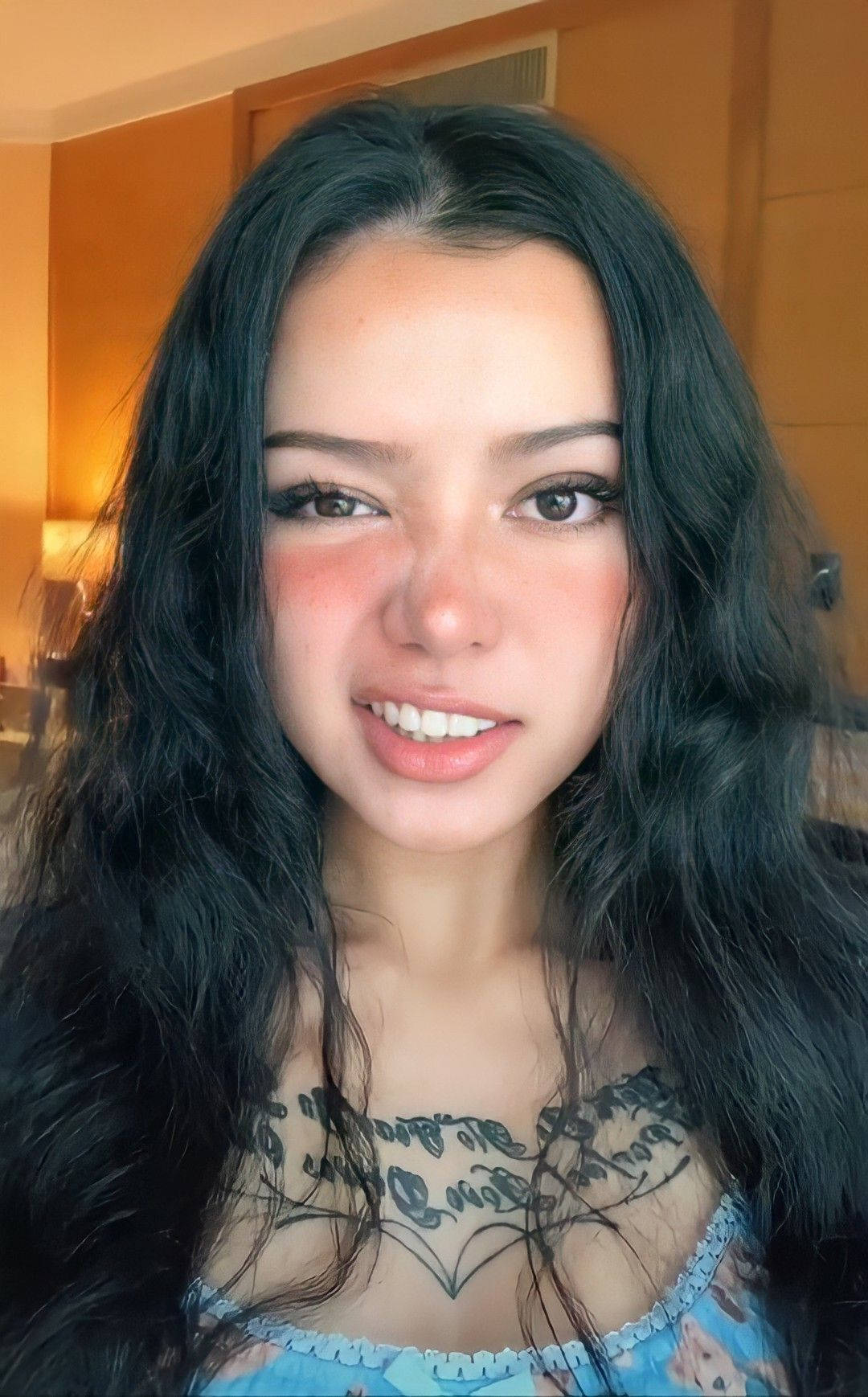 Bella Poarch 桌布 壁紙 — 一位擁有長長黑髮和胸口刺青的年輕女性，在溫暖的室內環境中露出溫暖的笑容。