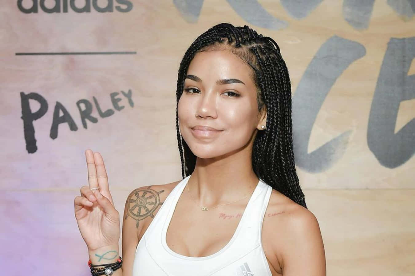 jhene aiko 桌布 壁紙 — 一位擁有長黑辮子和船輪紋身的女性在品牌活動中微笑比出和平手勢。
