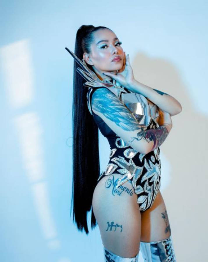 Bella Poarch 桌布 壁紙 — 一位擁有長黑髮和刺青的女性，在冷藍色燈光下，穿著金屬緊身衣，自信地擺姿勢，背景是純白牆壁。