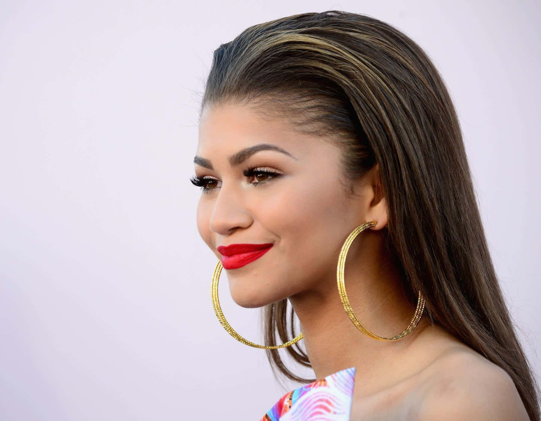 Zendaya 桌布 壁紙 — 一位擁有長黑髮和大膽紅唇的魅力女性，戴著大金圈耳環微笑。
