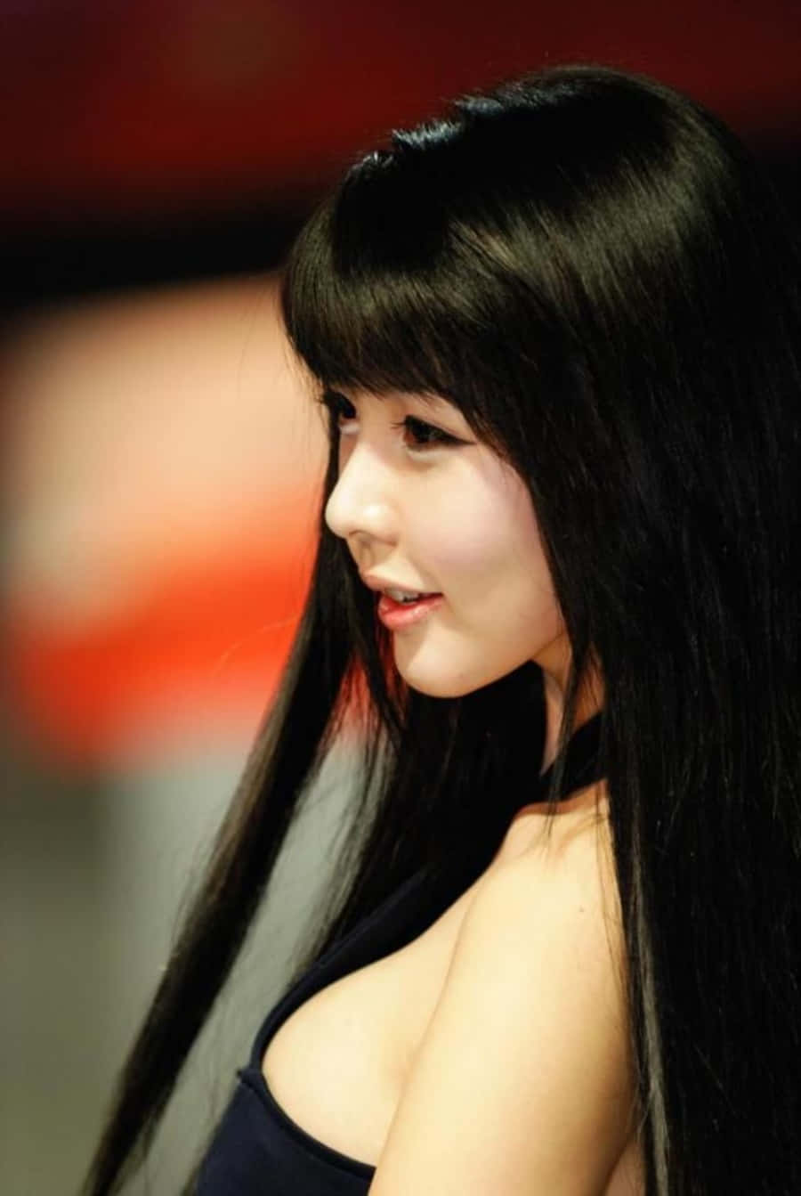 美女 桌布 壁紙 — 一位擁有長黑髮和瀏海的年輕女性，在昏暗的室內環境中，背景為模糊的紅色，溫柔地微笑。