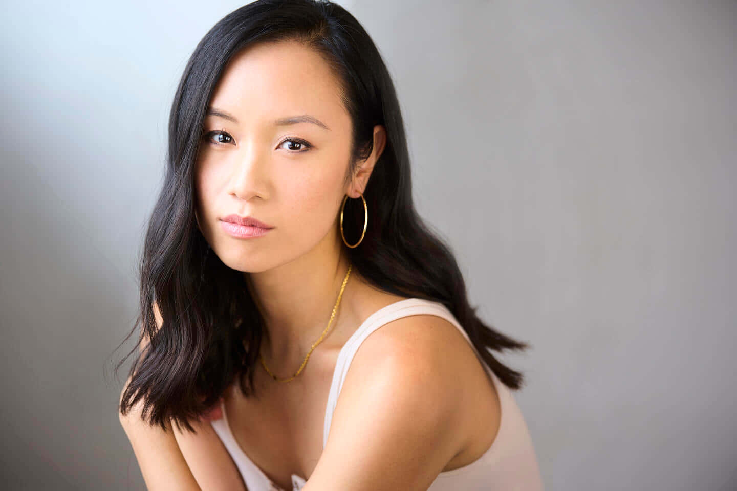 Ellen Wong 桌布 壁紙 — 一位擁有長黑髮和金色圓環耳環的女性，在灰色中性工作室背景下柔和地凝視相機。