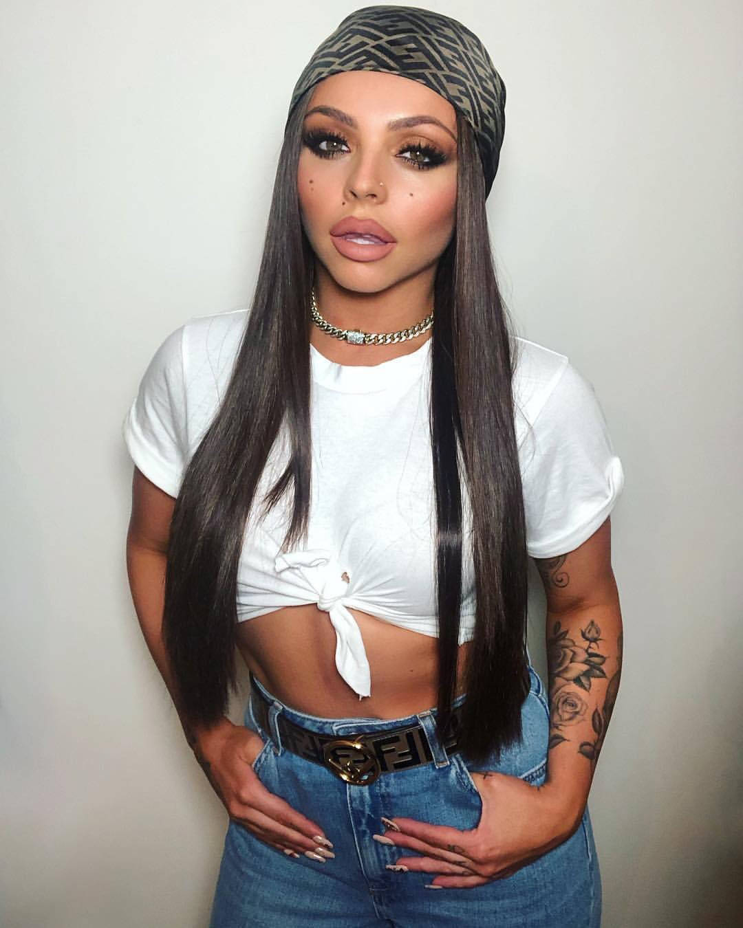 Jesy Nelson 桌布 壁紙 — 一位擁有長黑髮和鮮豔妝容的女性，穿著白色綁帶短版上衣和牛仔褲，自信地靠在素牆上擺姿勢。