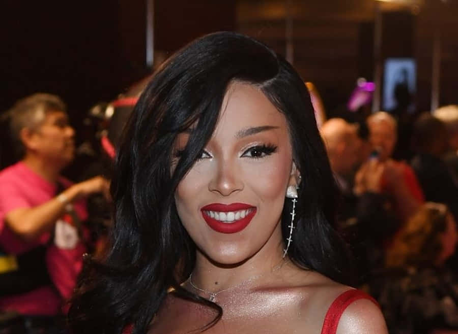 Doja Cat 桌布 壁紙 — 一位擁有長黑髮和鮮豔紅唇的魅力女性，在溫暖燈光下於擁擠的活動中自信微笑。