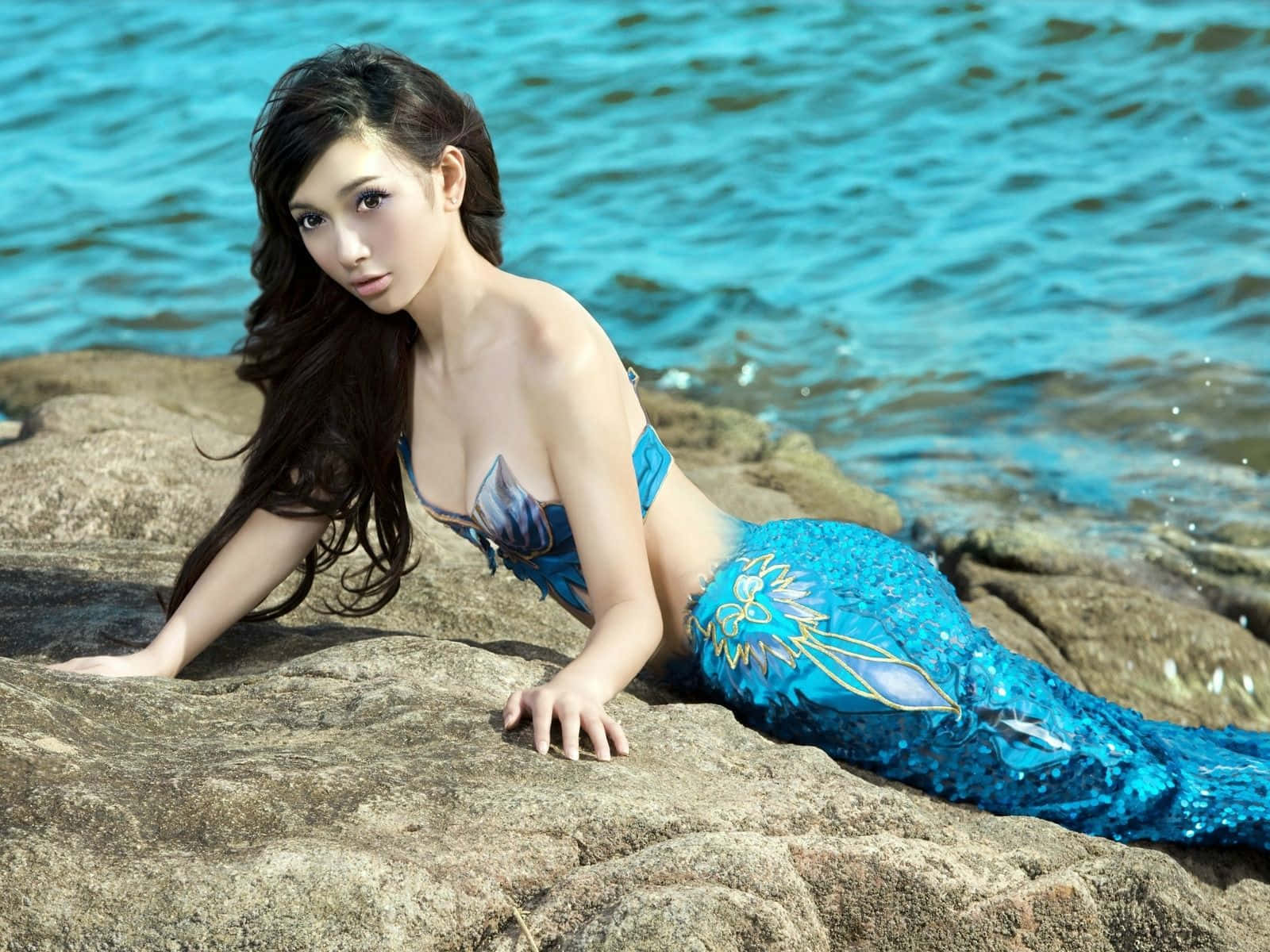 Real Mermaid 桌布 壁紙 — 一位擁有閃亮藍色美人魚尾巴的女性，在陽光普照的岩石上倚靠著，旁邊是碧綠色的海水。