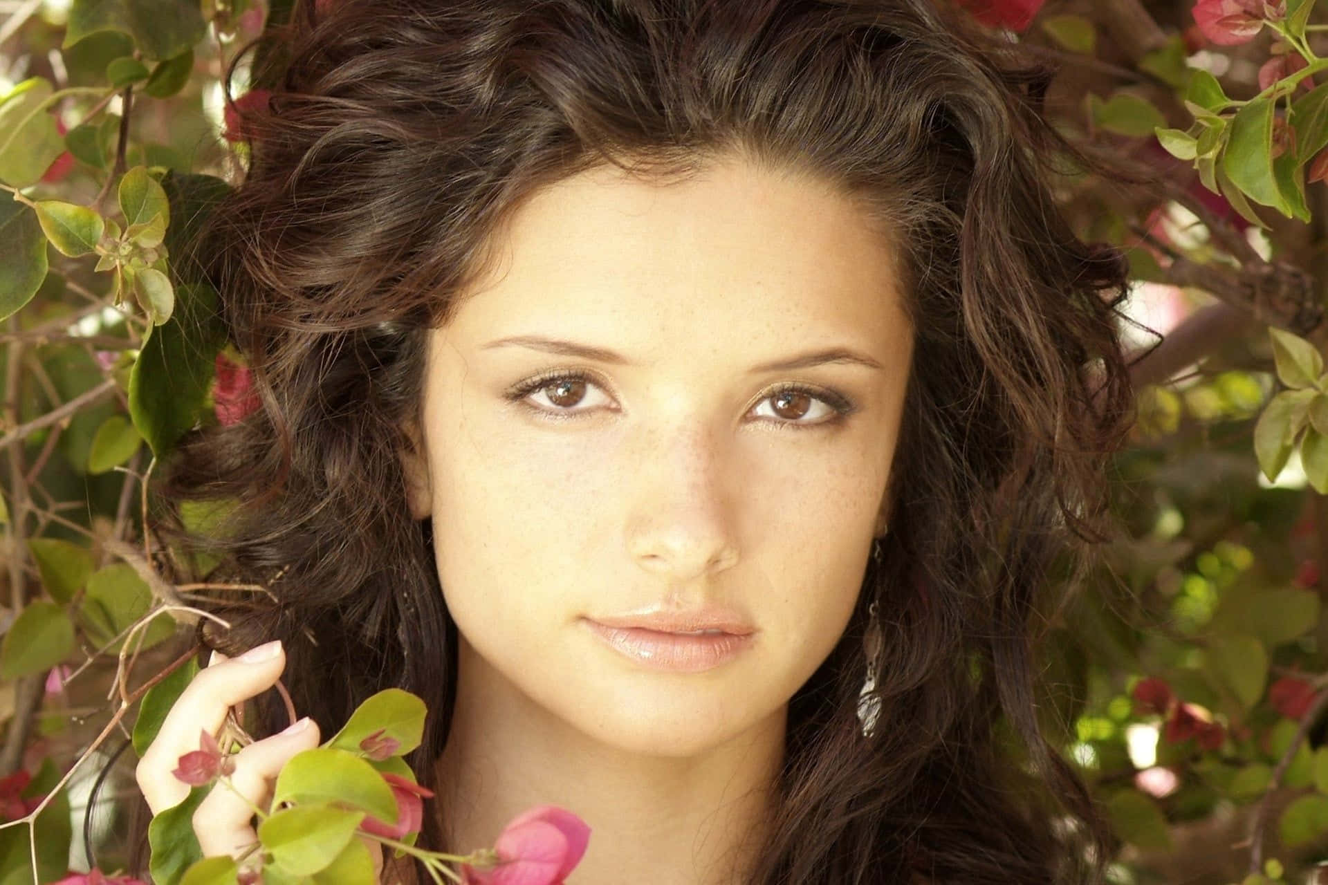 Alice Greczyn 桌布 壁紙 — 一位擁有雀斑和深色捲髮的年輕女性，在充滿活力的綠色樹葉和粉紅色花朵中溫柔地凝視。