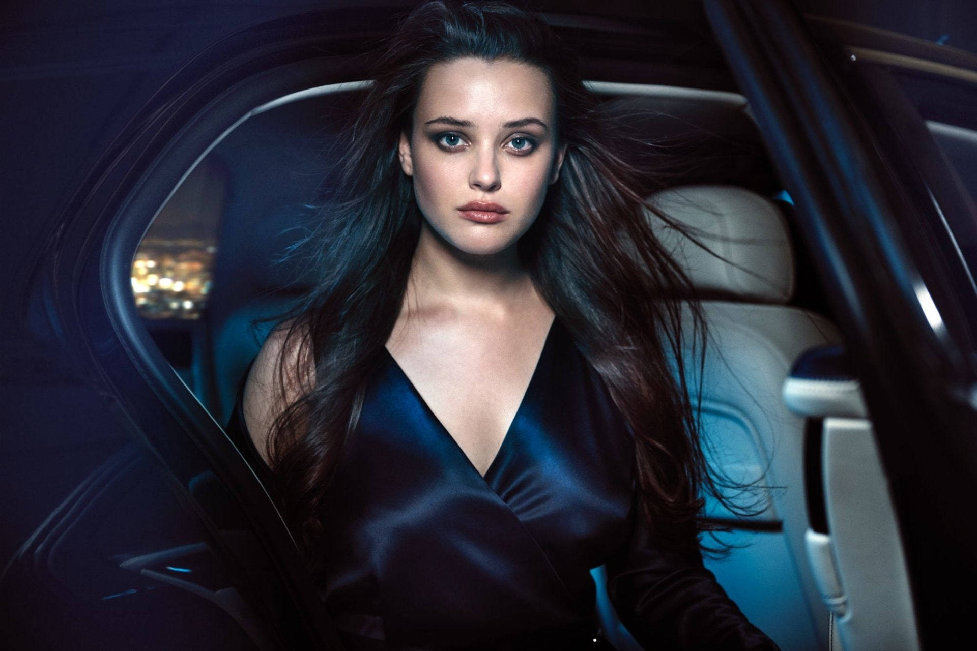 Katherine Langford 桌布 壁紙 — 一位擁有飄逸深色長髮的女性夜晚坐在車內，穿著時尚的黑色緞面連衣裙，被身後的都市燈光照亮。