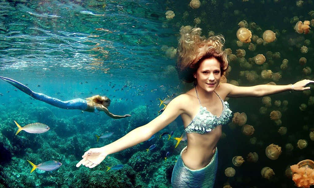 Mermaid Real Life 桌布 壁紙 — 一位擁有飄逸紅髮的美人魚在珊瑚附近的水中微笑游泳，周圍有發光的水母和色彩繽紛的魚類。