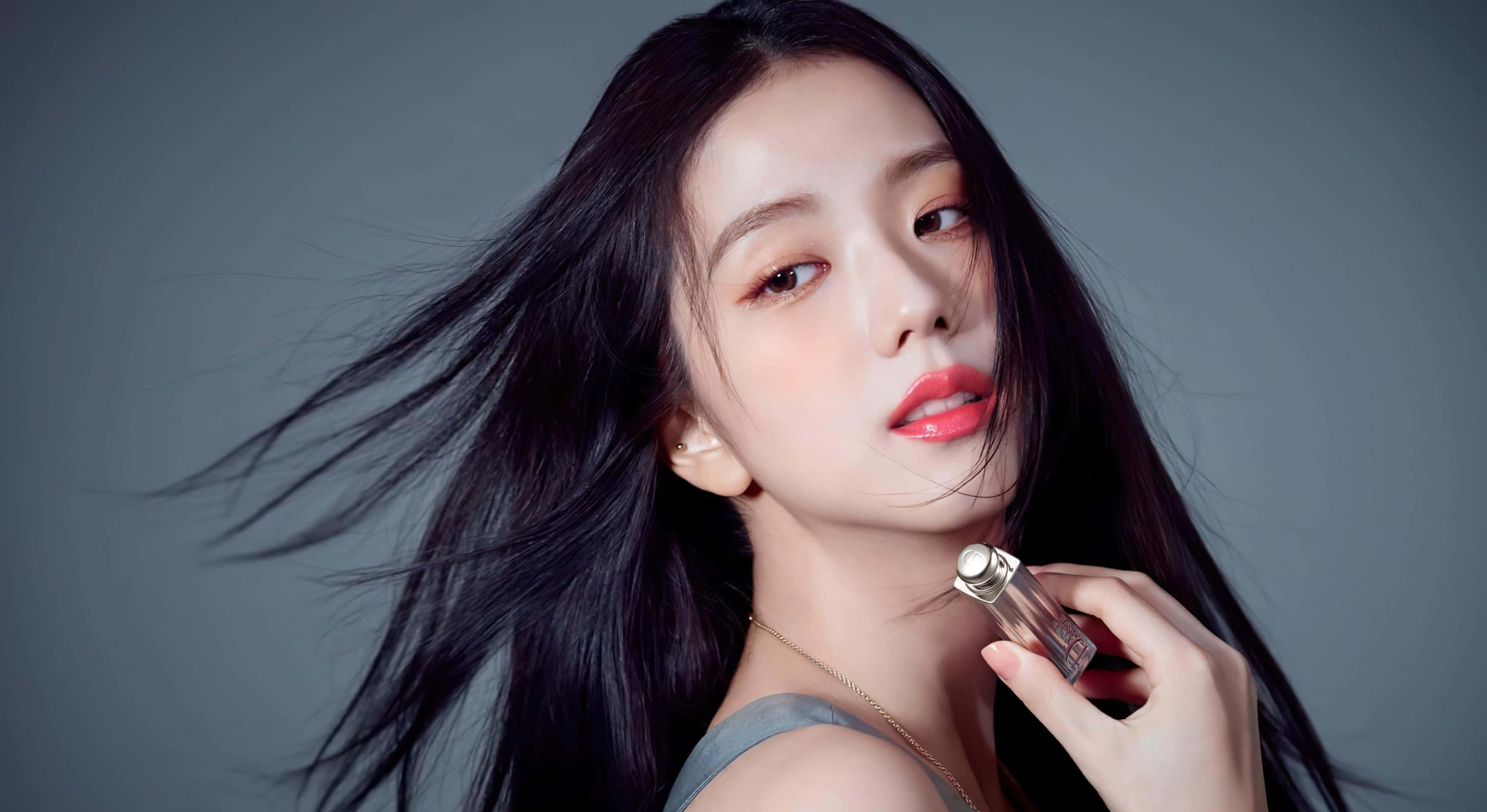 BLACKPINK 桌布 壁紙 — 一位擁有飄逸黑髮和紅唇的女性，在柔和的灰色背景下拿著香水瓶。
