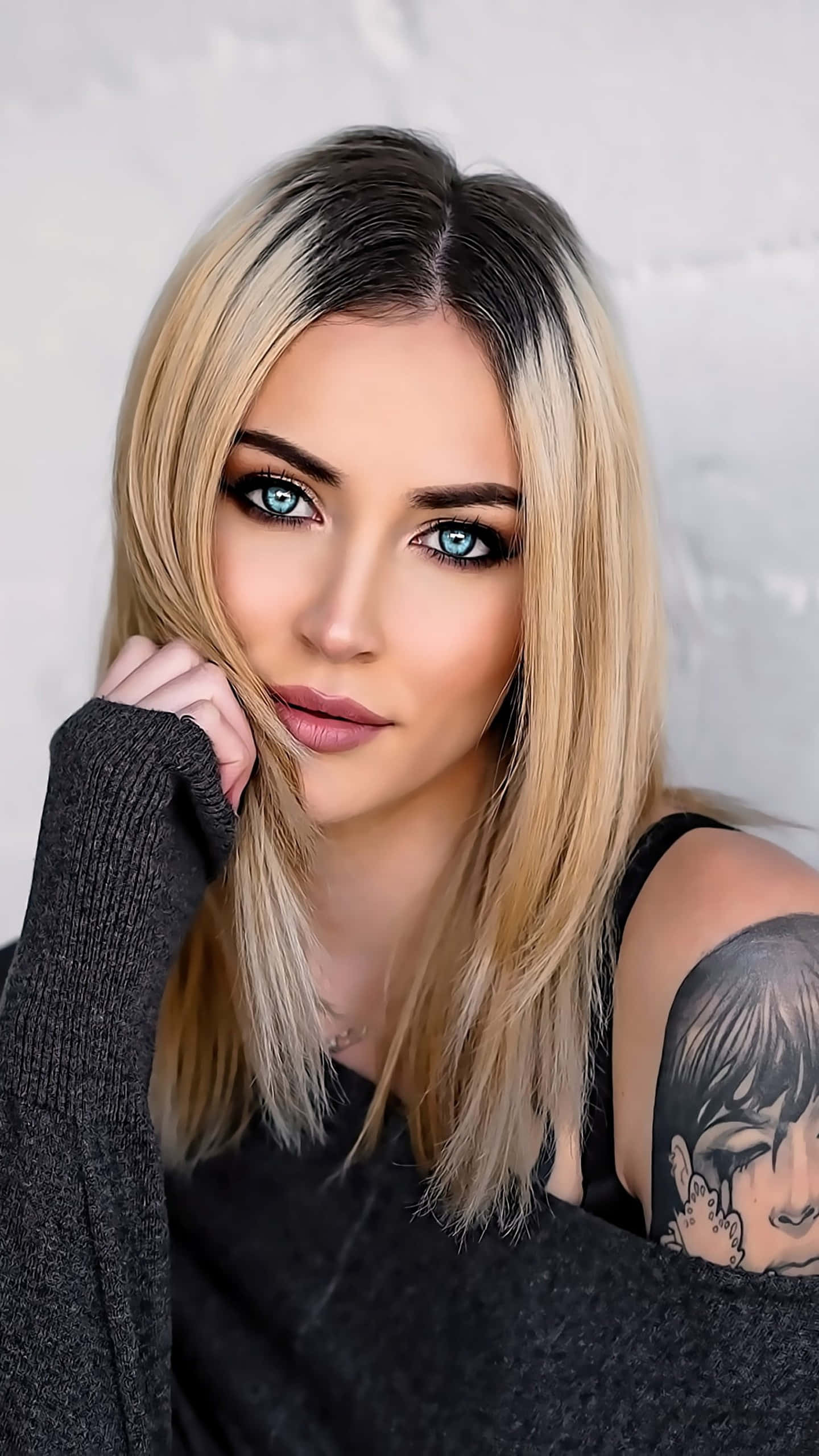 Tattoo Girl 桌布 壁紙 — 一位擁有驚豔藍眼睛和金色頭髮的女性，將下巴擱在手上，靠著紋理白色牆壁。