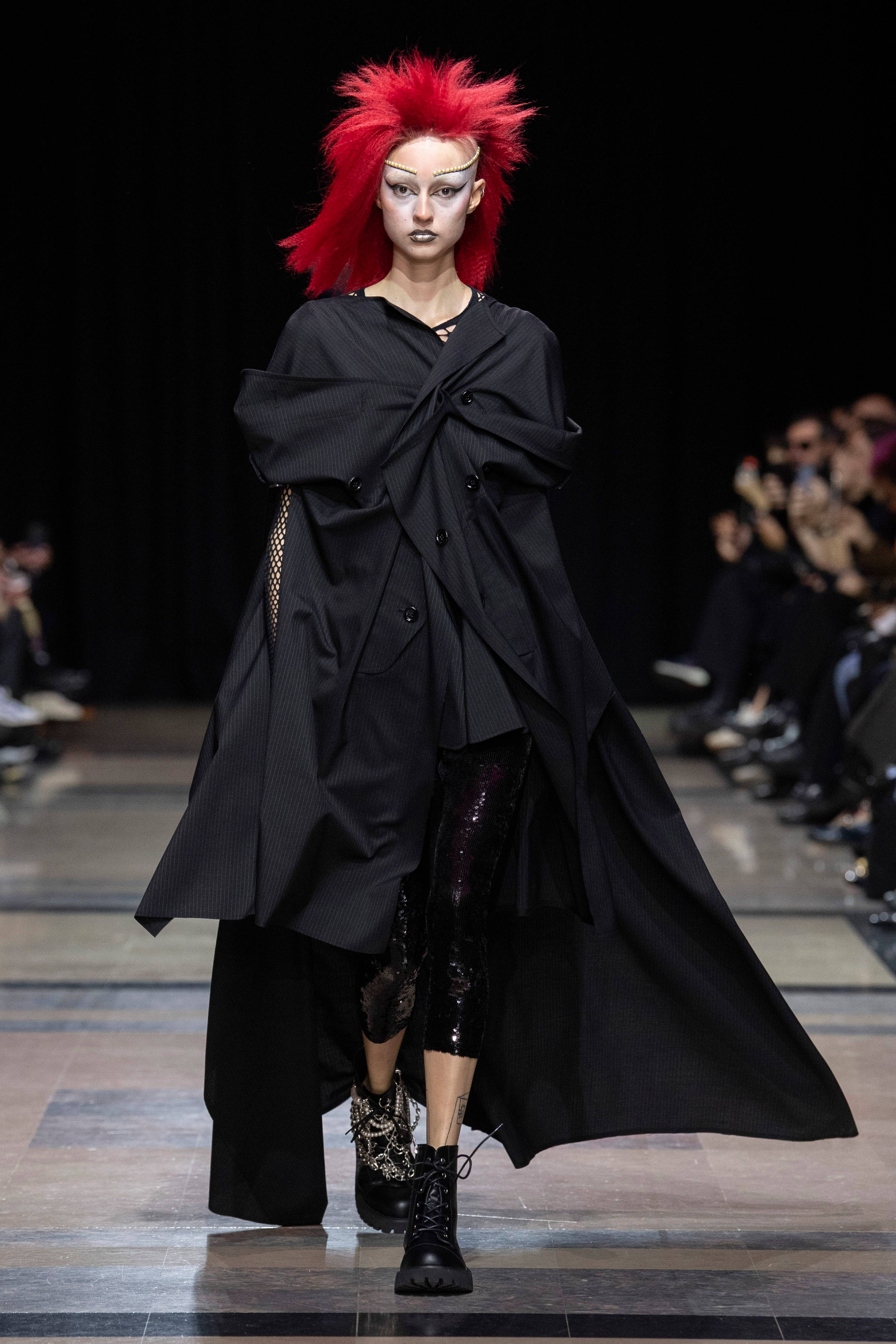 Clothes 桌布 壁紙 — 一位擁有鮮紅色尖刺頭髮和大膽妝容的模特兒，自信地穿著一件戲劇性的超大黑色外套和閃亮緊身褲走在天橋上。
