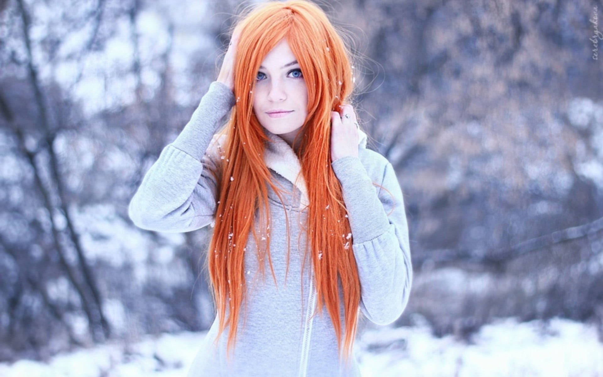 Cute Winter 桌布 壁紙 — 一位擁有鮮豔橙色頭髮的年輕女性站在雪地森林中，穿著灰色連帽衫，雪花輕輕飄落在她周圍。