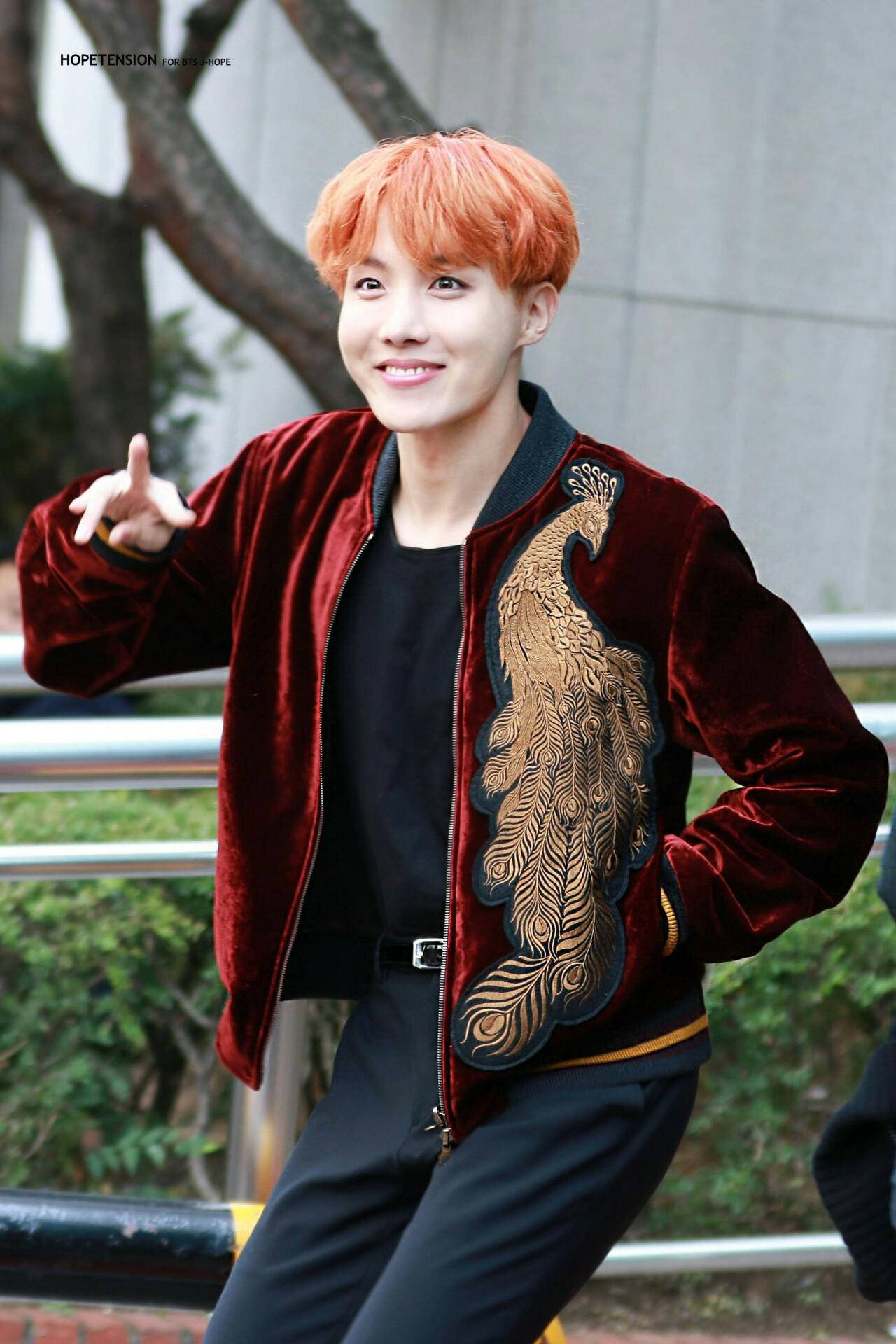 jhope 桌布 壁紙 — 一位擁有鮮豔橙色頭髮的年輕男子，穿著一件飾有精緻金色孔雀刺繡的濃郁紅色天鵝絨外套，露出燦爛的笑容。