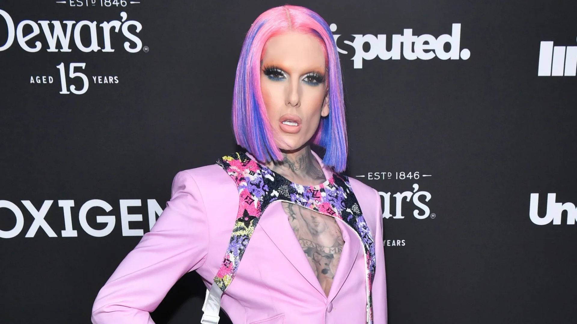 Jeffree Star 桌布 壁紙 — 一位擁有鮮豔粉紅和紫色頭髮及刺青的人，在品牌活動中穿著帶有花卉肩帶的粉紅色西裝外套。