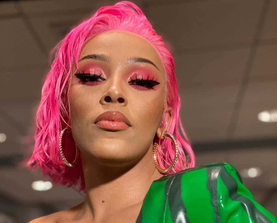 Doja Cat 桌布 壁紙 — 一位擁有鮮豔粉紅色頭髮和大膽妝容的女性，穿著綠色和黑色的服裝，戴著金色圓環耳環。
