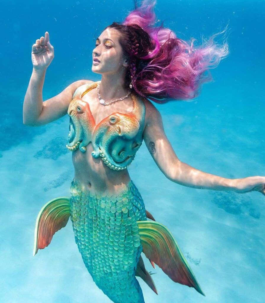 美人魚 桌布 壁紙 — 一位擁有鮮豔粉紅色頭髮和色彩繽紛的章魚主題上衣的美人魚，在清澈的藍色水中優雅地滑行。