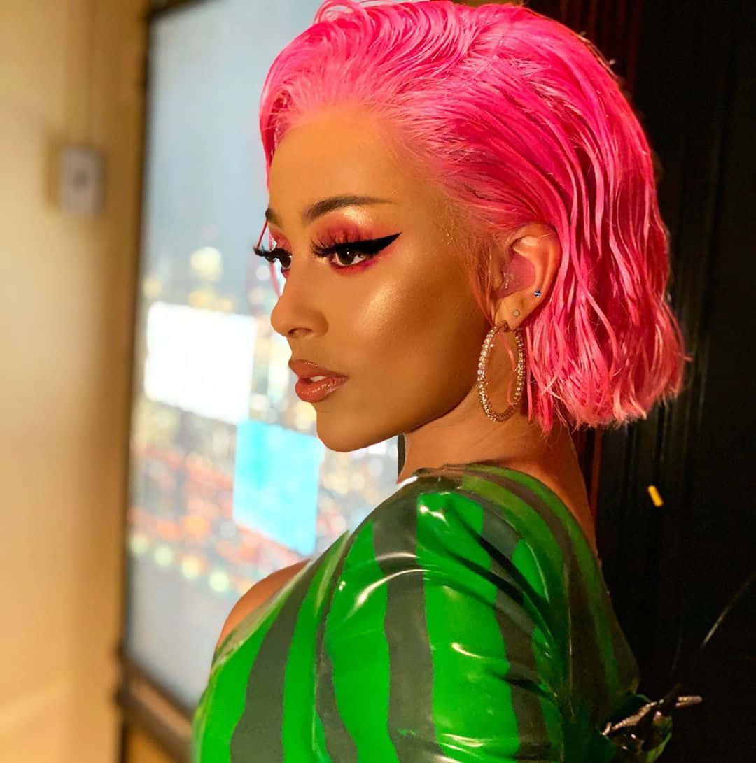 Doja Cat 桌布 壁紙 — 一位擁有鮮豔粉紅頭髮和濃妝的女性，穿著閃亮的綠色條紋上衣，凝視窗外。