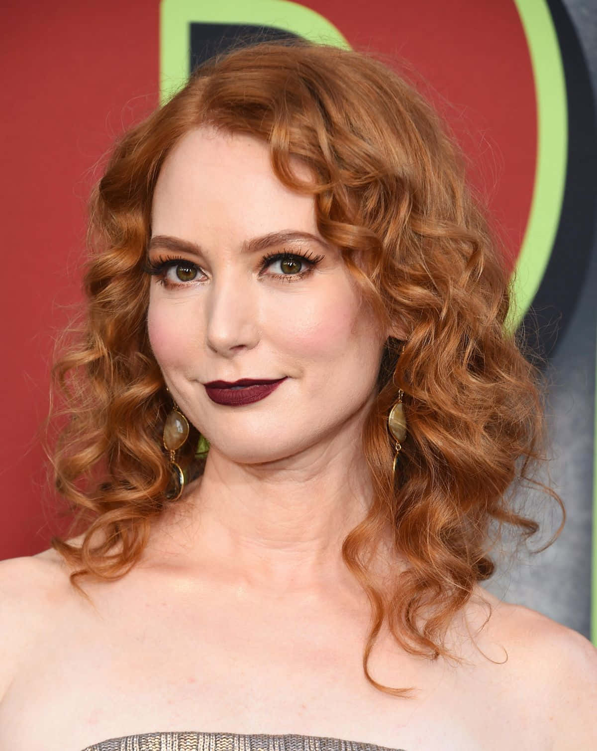 Alicia Witt 桌布 壁紙 — 一位擁有鮮豔紅色捲髮和濃烈深色唇膏的女性，在色彩繽紛的活動背景下溫柔微笑。