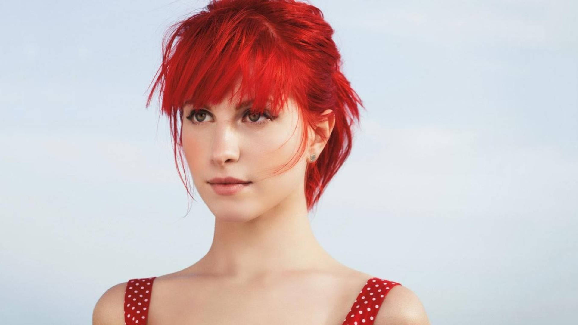 Hayley Williams 桌布 壁紙 — 一位擁有鮮豔紅髮和紅色圓點上衣的女性，在柔和的藍天背景下平靜地凝視。