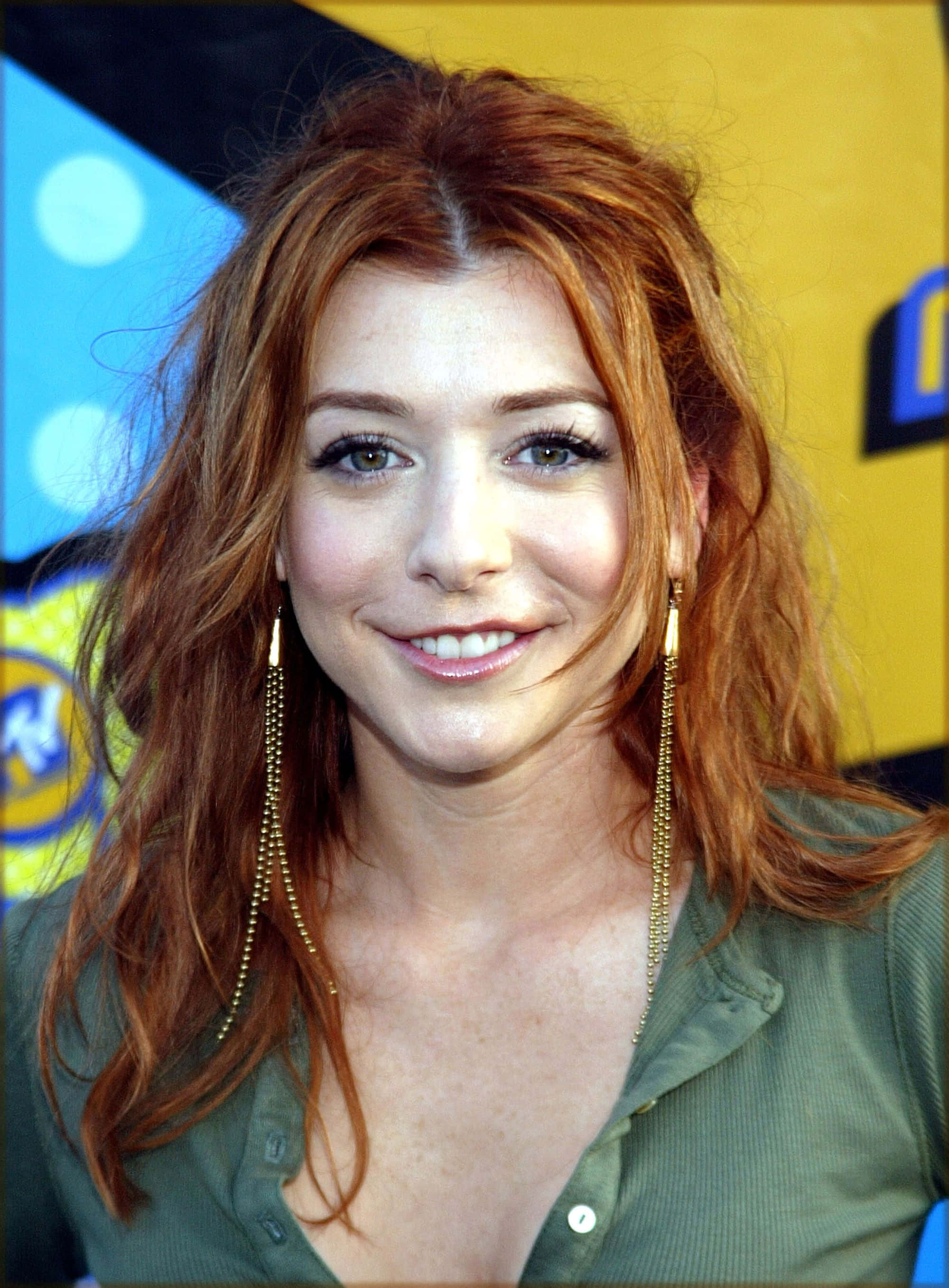 Alyson Hannigan 桌布 壁紙 — 一位擁有鮮豔紅髮和金色垂墜耳環的女性，在色彩繽紛的黃藍背景下溫暖微笑。