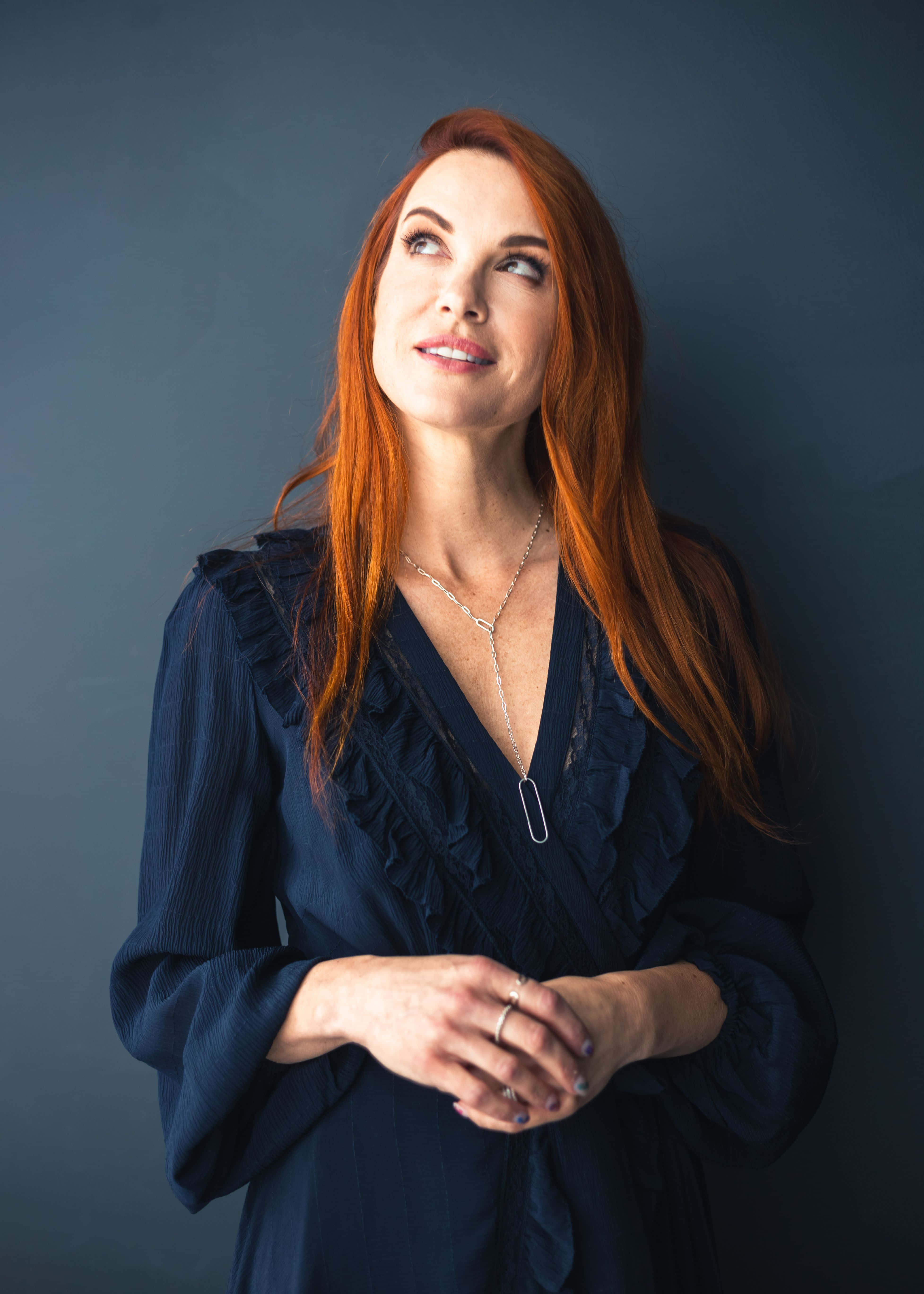 Danneel Ackles 桌布 壁紙 — 一位擁有鮮豔紅髮的女性，身穿深藍色連衣裙，在柔和的灰色背景下向上凝視。
