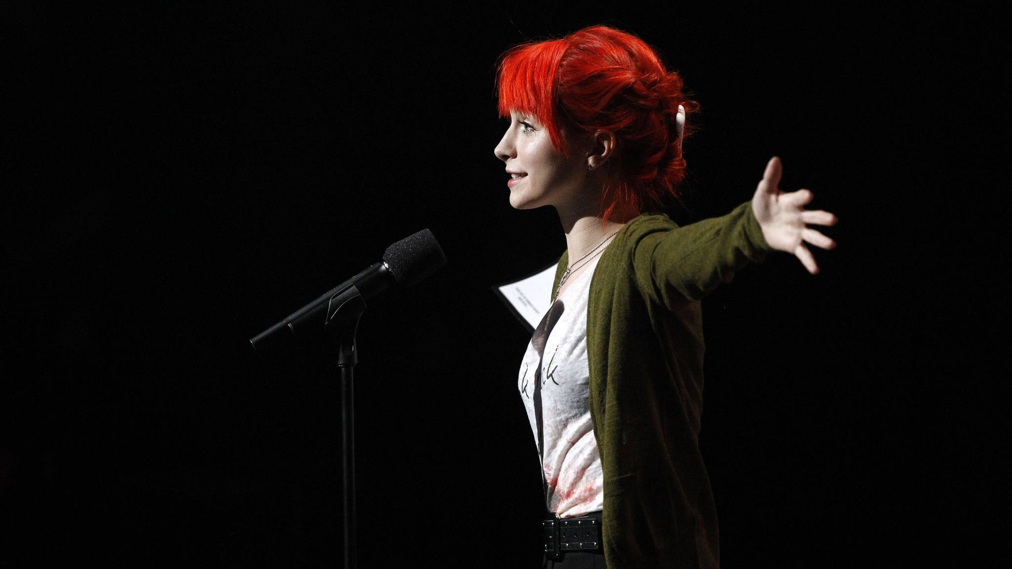 Hayley Williams 桌布 壁紙 — 一位擁有鮮豔紅髮的女性在戲劇性的聚光燈下在舞台上表演，在黑色背景前在麥克風旁邊富有表現力地做手勢。