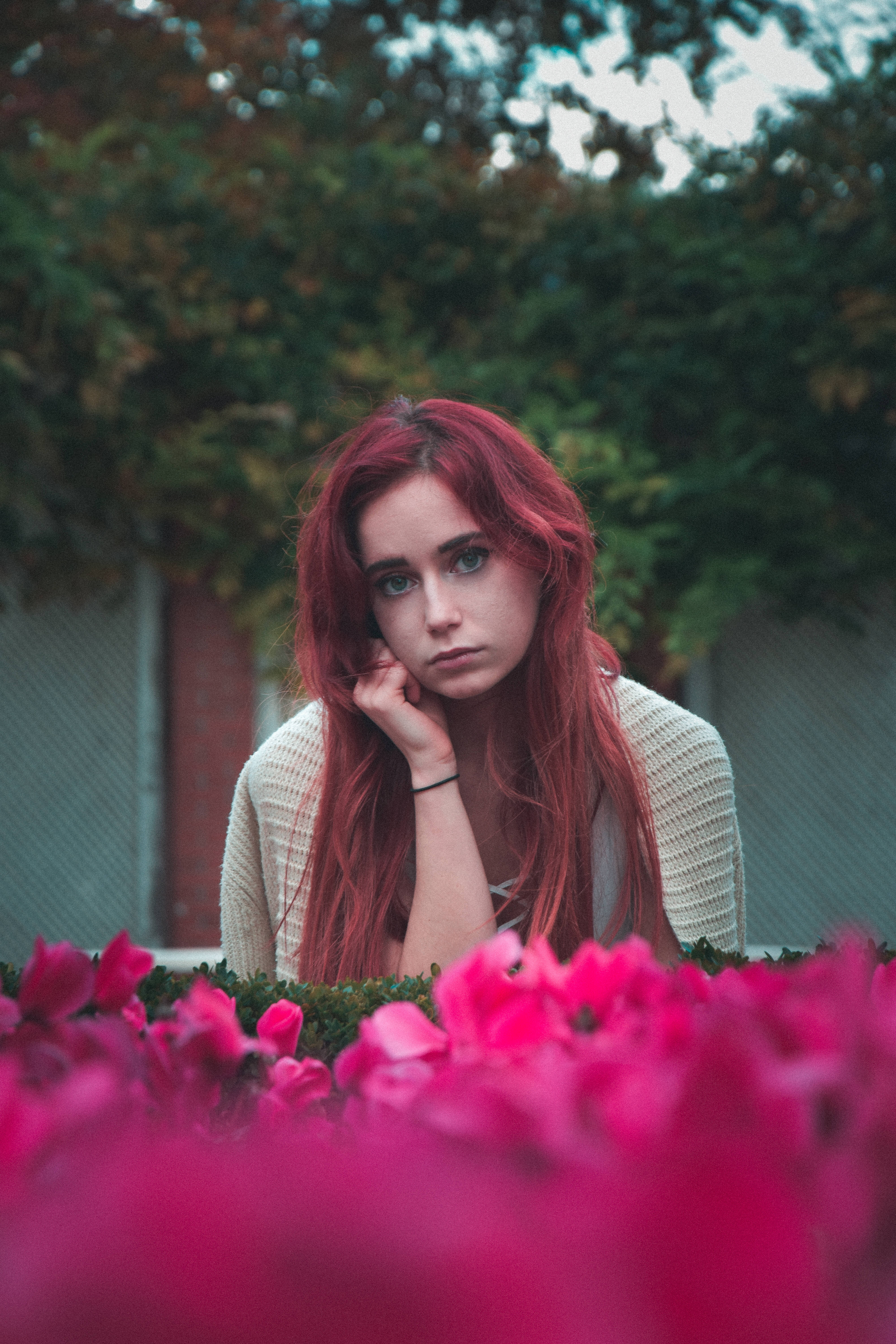 Cool Sad 桌布 壁紙 — 一位擁有鮮豔紅髮的年輕女子，在柔和模糊的花園環境中，若有所思地凝視著一片鮮豔的粉紅色花朵。