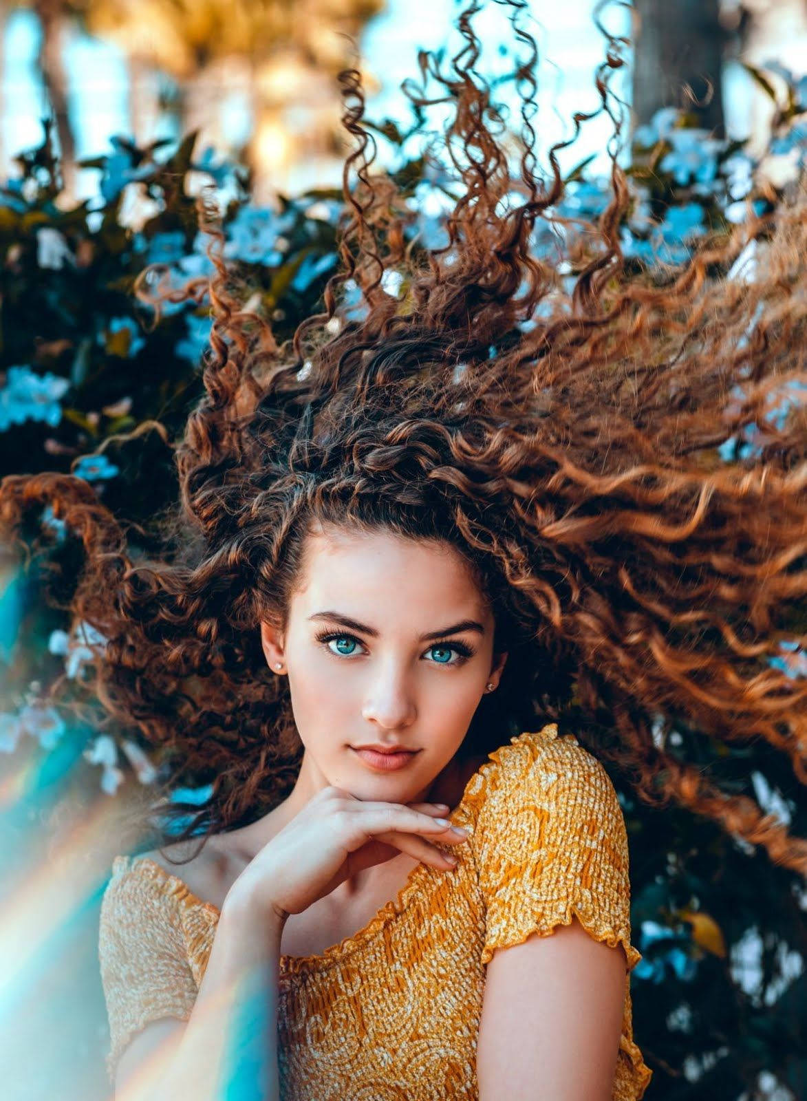 hair flip 桌布 壁紙 — 一位擁有鮮豔藍色眼睛和飄逸棕色捲髮的女性，在藍色花朵和柔和陽光的背景下，穿著金色質感上衣。