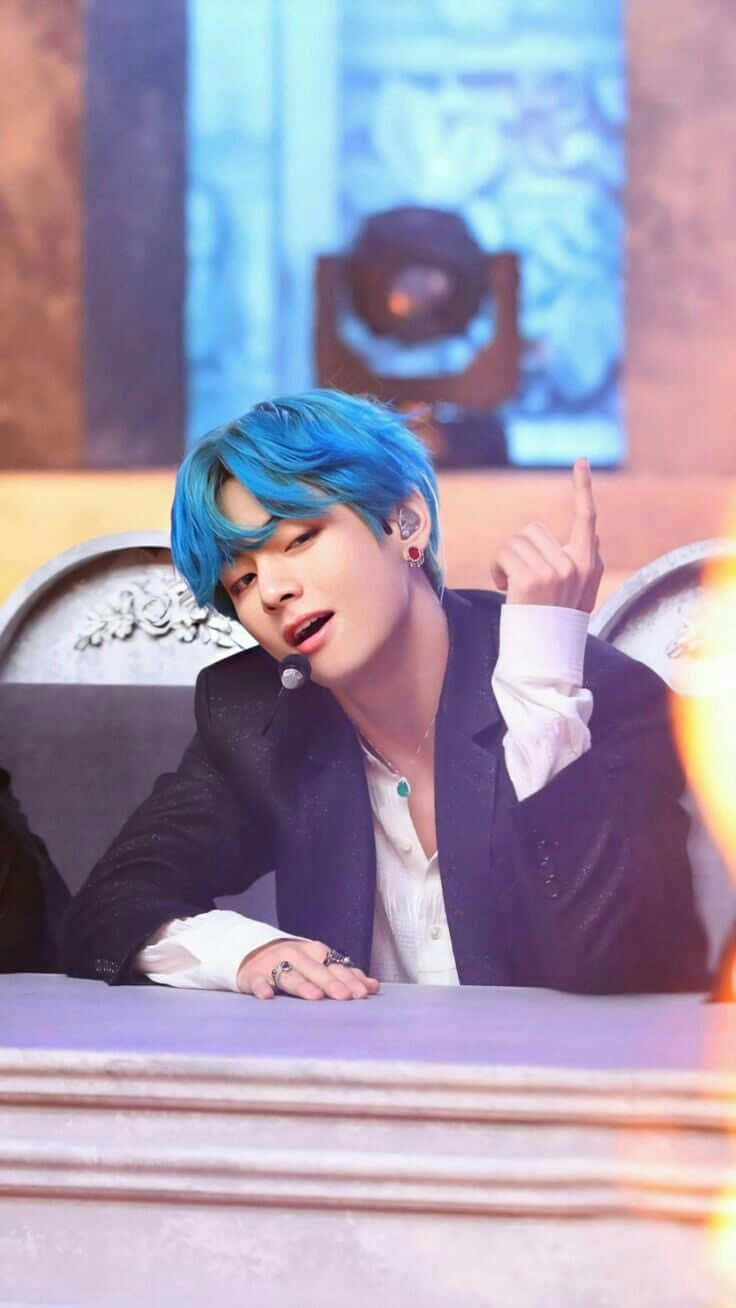 K-pop 桌布 壁紙 — 一位擁有鮮豔藍色頭髮的年輕男子，在溫暖的舞台燈光下坐在舞台桌前做手勢。