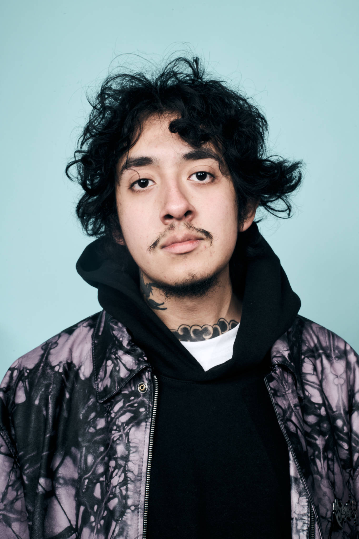 Cuco 桌布 壁紙 — 一位擁有黑色捲髮和頸部刺青的年輕男子，在純淺藍色背景下直視鏡頭。