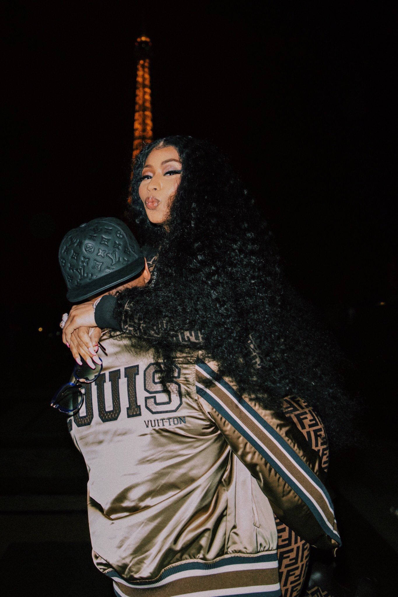 Nicki 桌布 壁紙 — 一位擁有黑色長曲髮的女性在夜晚擁抱著一位穿著金色Louis Vuitton外套的人，背景是發光的艾菲爾鐵塔。